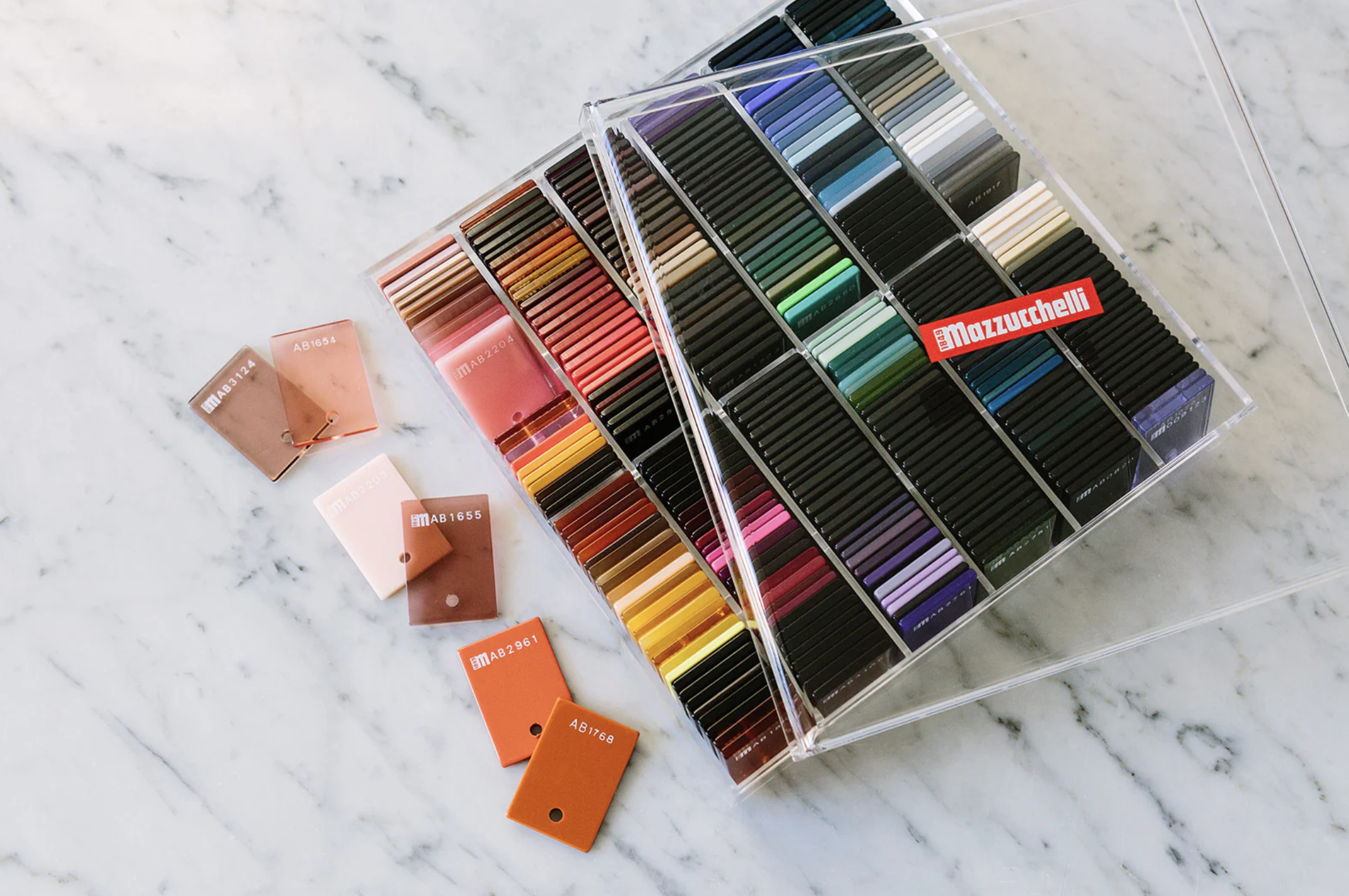A clear plastic storage box containing numerous colored markers arranged by color on a white marble surface. Several color swatches with labels are placed in front of the box.