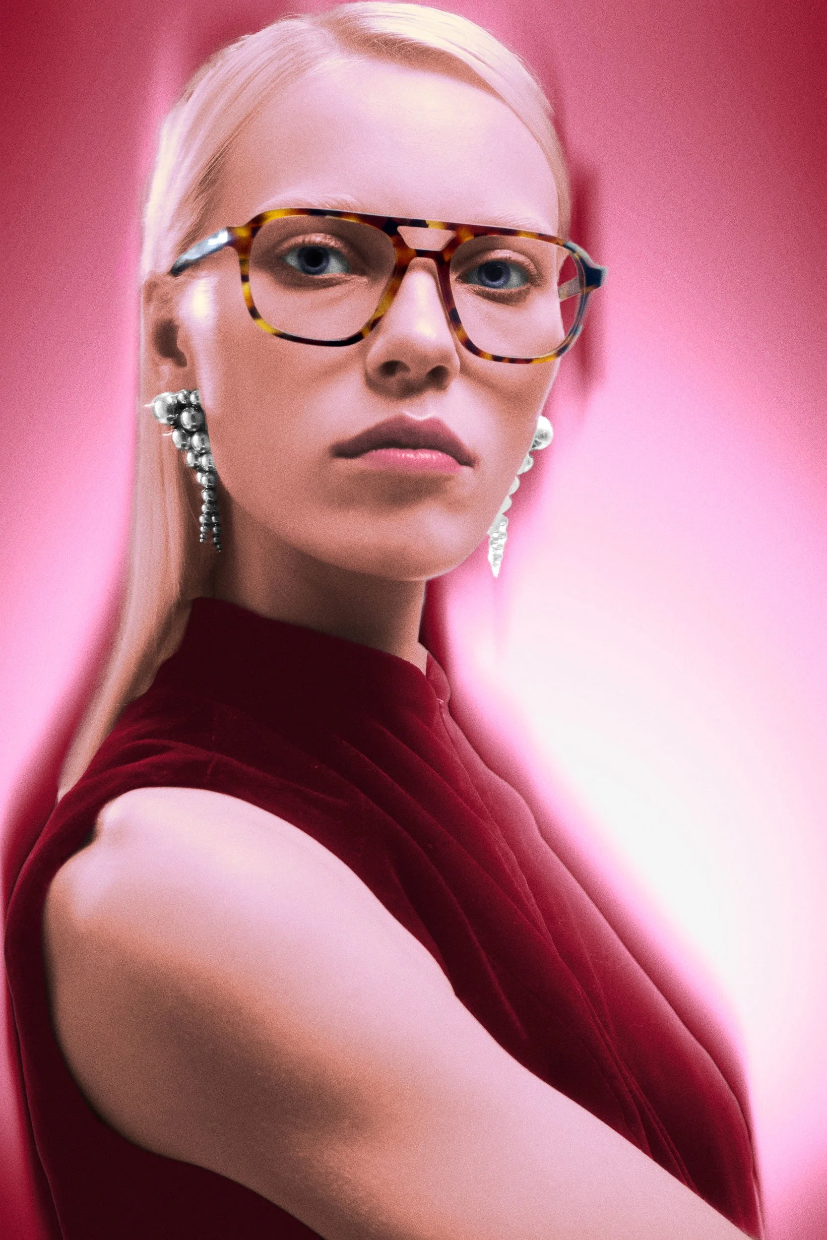 Close-up of a woman with platinum blonde hair, blue eyes, and pierced ears wearing pearl earrings. She is wearing green tortoiseshell glasses and a deep red velvet dress against a pink background.