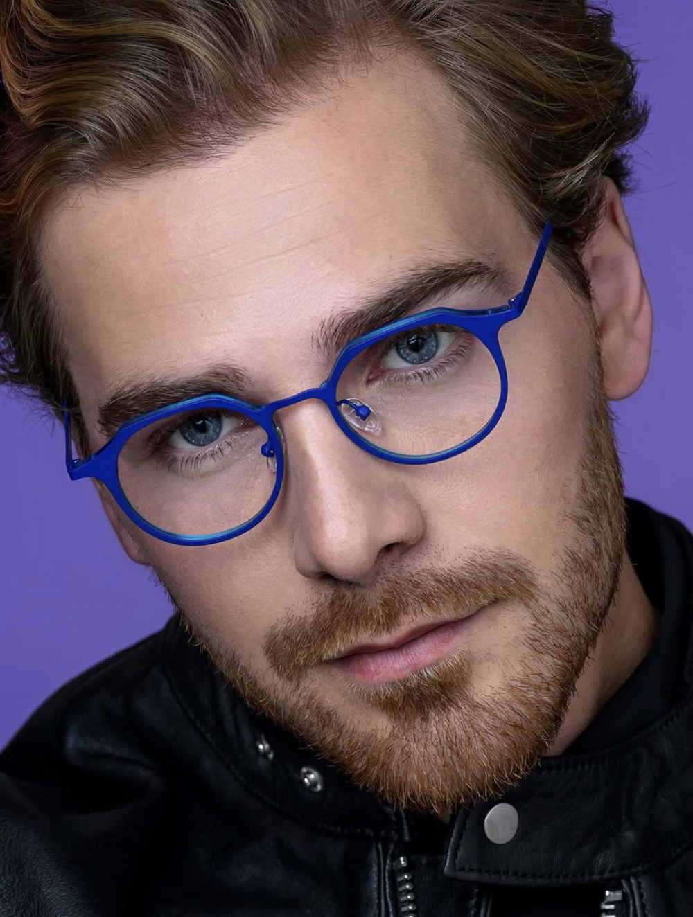 A close-up portrait of a young man with blue eyes, red hair, and a red beard, wearing blue glasses and a black leather jacket against a purple background.