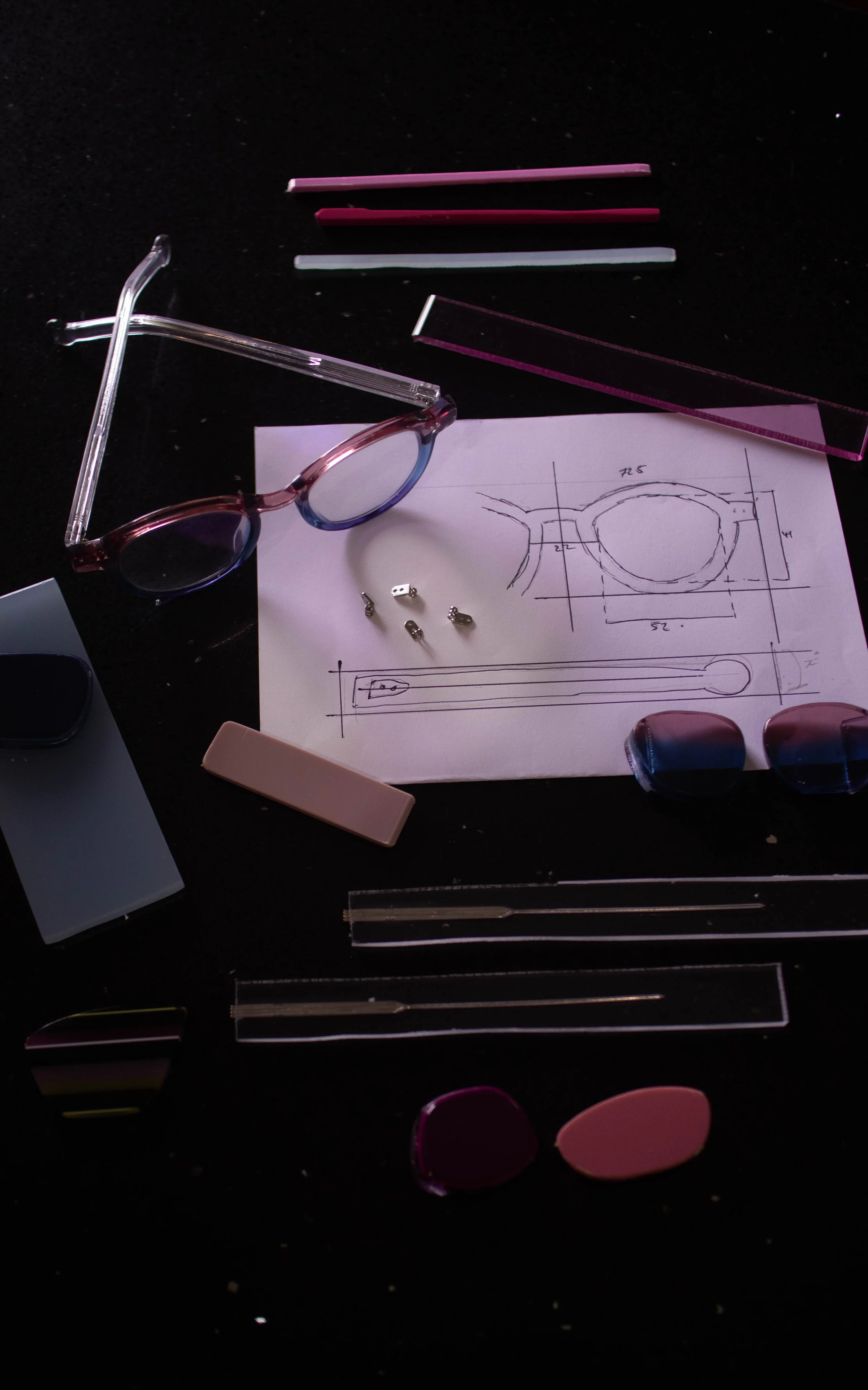 A workspace with medical or optical tools, including eyeglasses, lenses, a sketch of a frame, and various small parts and tools on a dark surface.