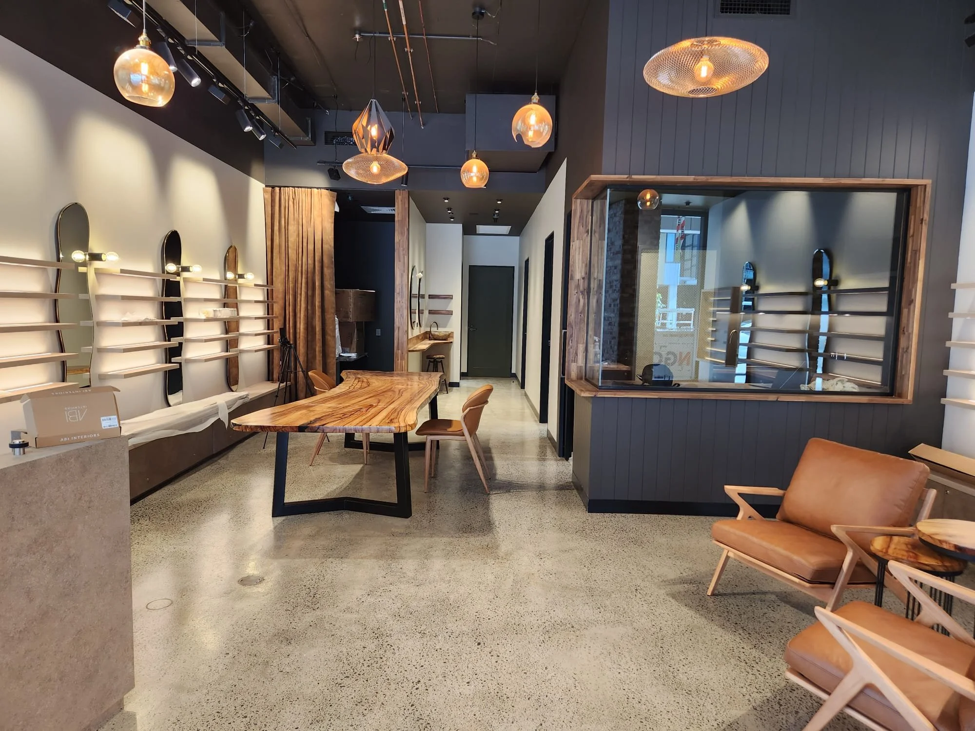 Interior of a modern cafe or lounge with wooden chairs and tables, pendant lights hanging from a dark ceiling, a large mirror on the wall reflecting natural light, and curtains dividing the space.