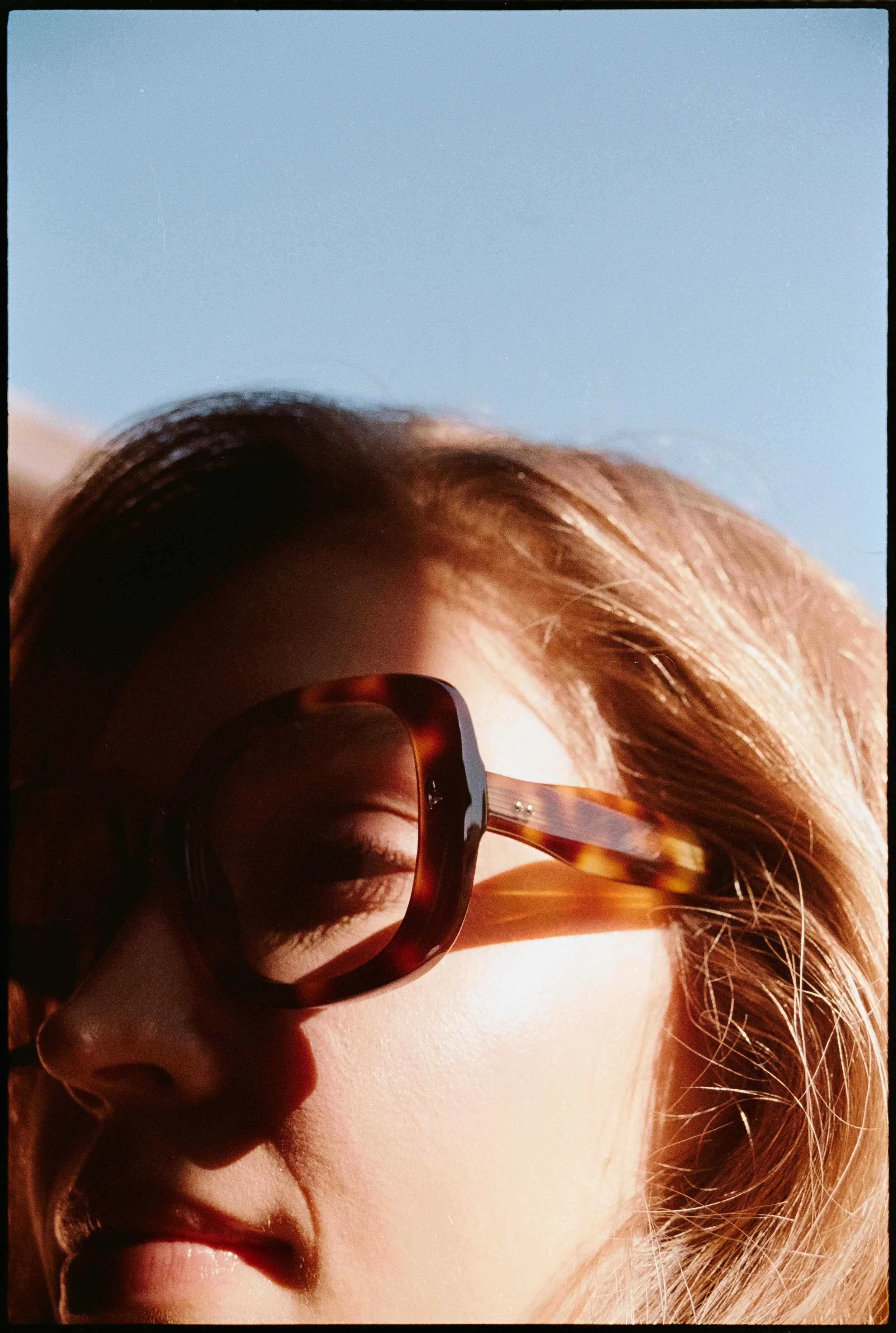 Close-up of a woman's face with sunglasses, light brown hair, and smooth skin, looking downward, against a clear blue sky.