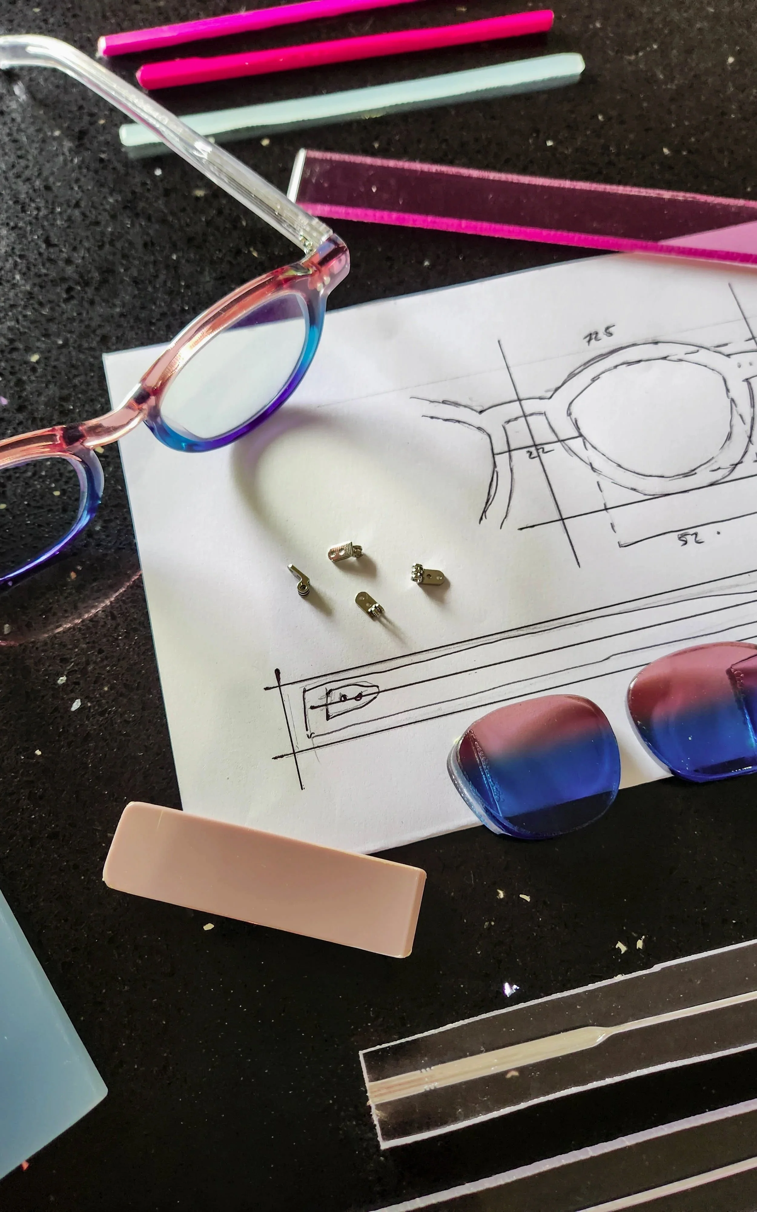 A pair of pink and blue glasses, several colorful sample glasses, a ruler, a sketch of an eyeglass frame on paper, small metal hinge parts, and a rectangular pink object on a black surface.