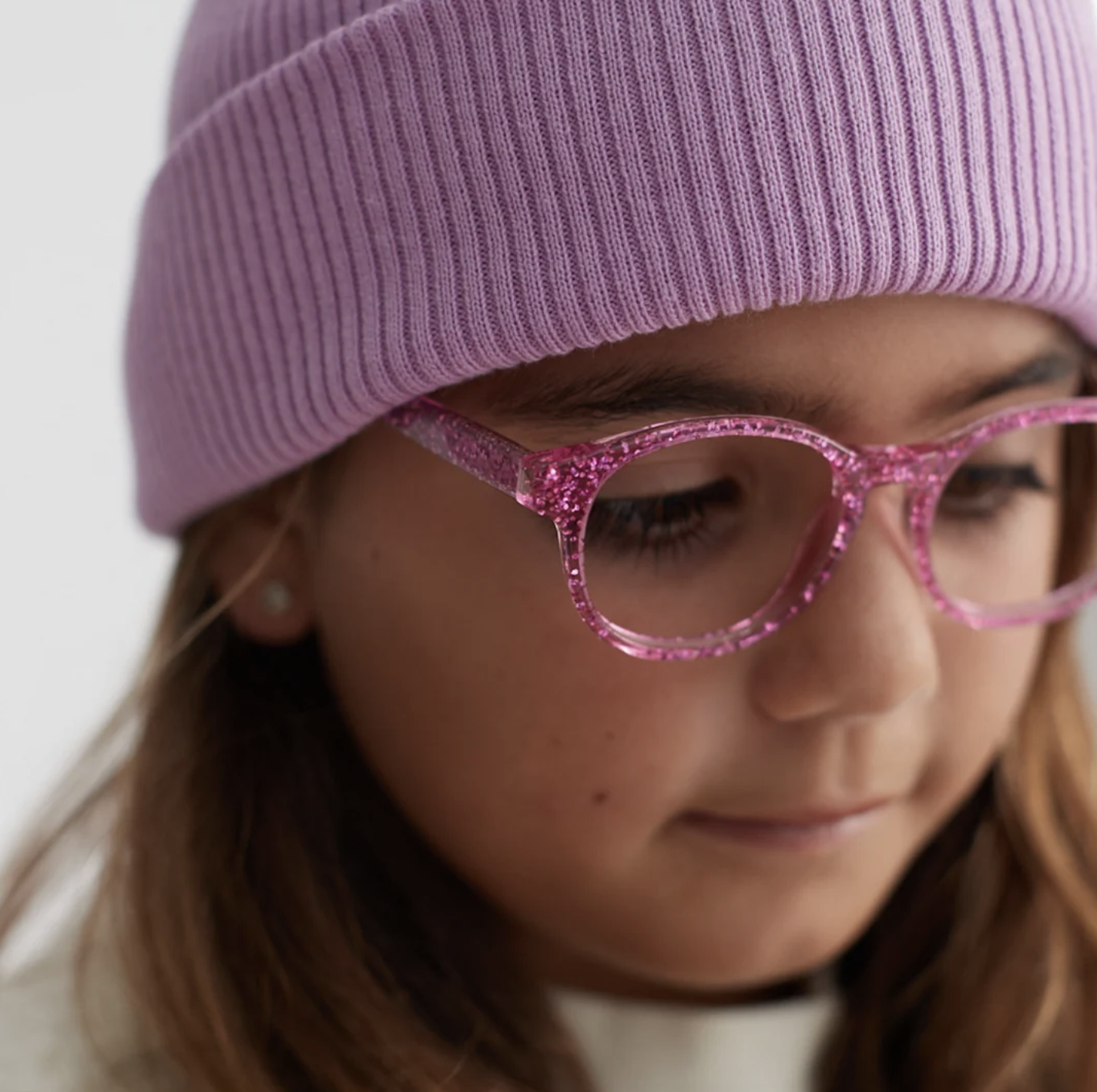 Close-up of a young woman wearing pink glittery glasses, a purple knit beanie, and with long brown hair.
