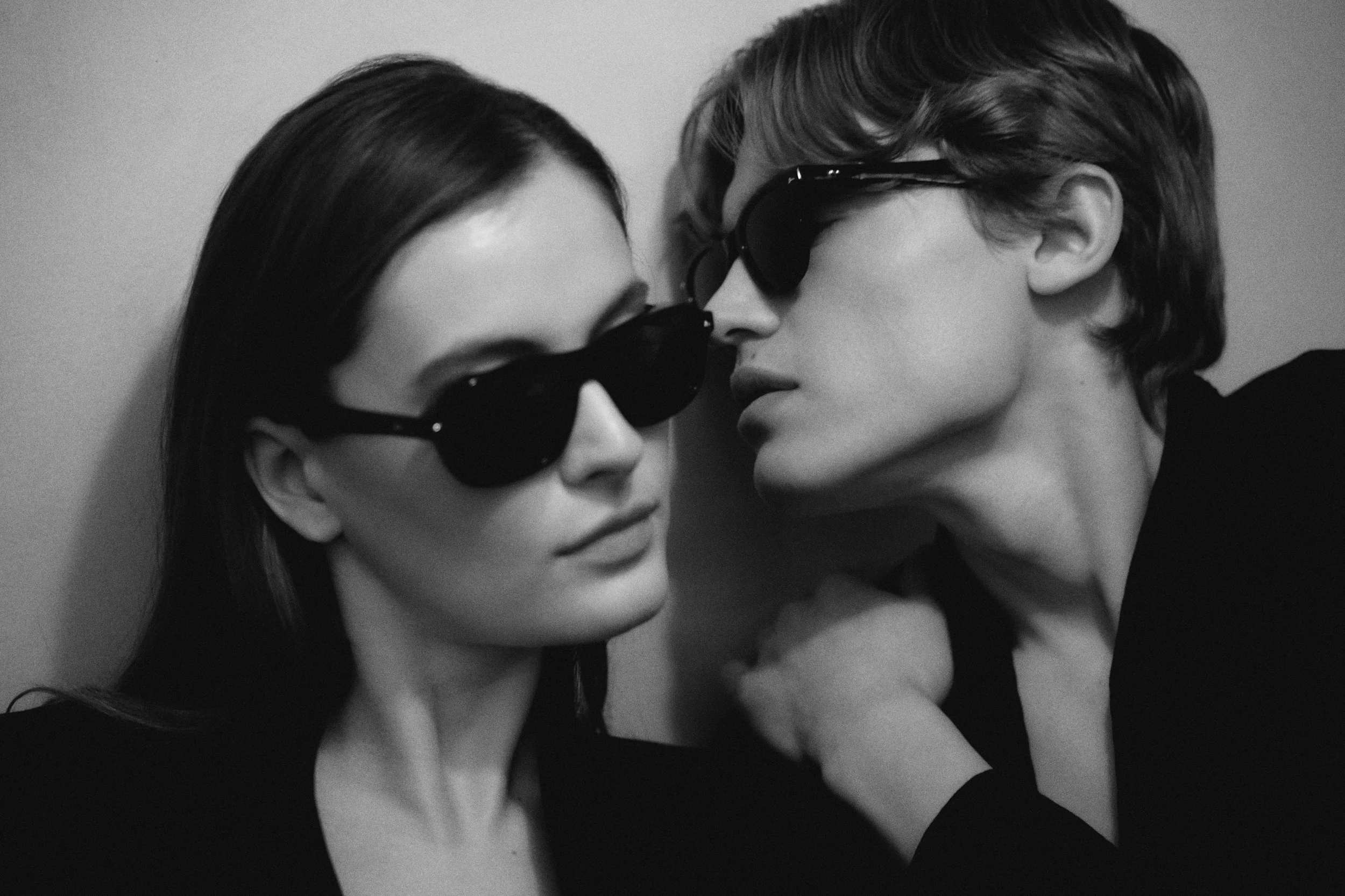 Two women wearing black sunglasses face close to each other, with their foreheads almost touching, in a black-and-white photo.