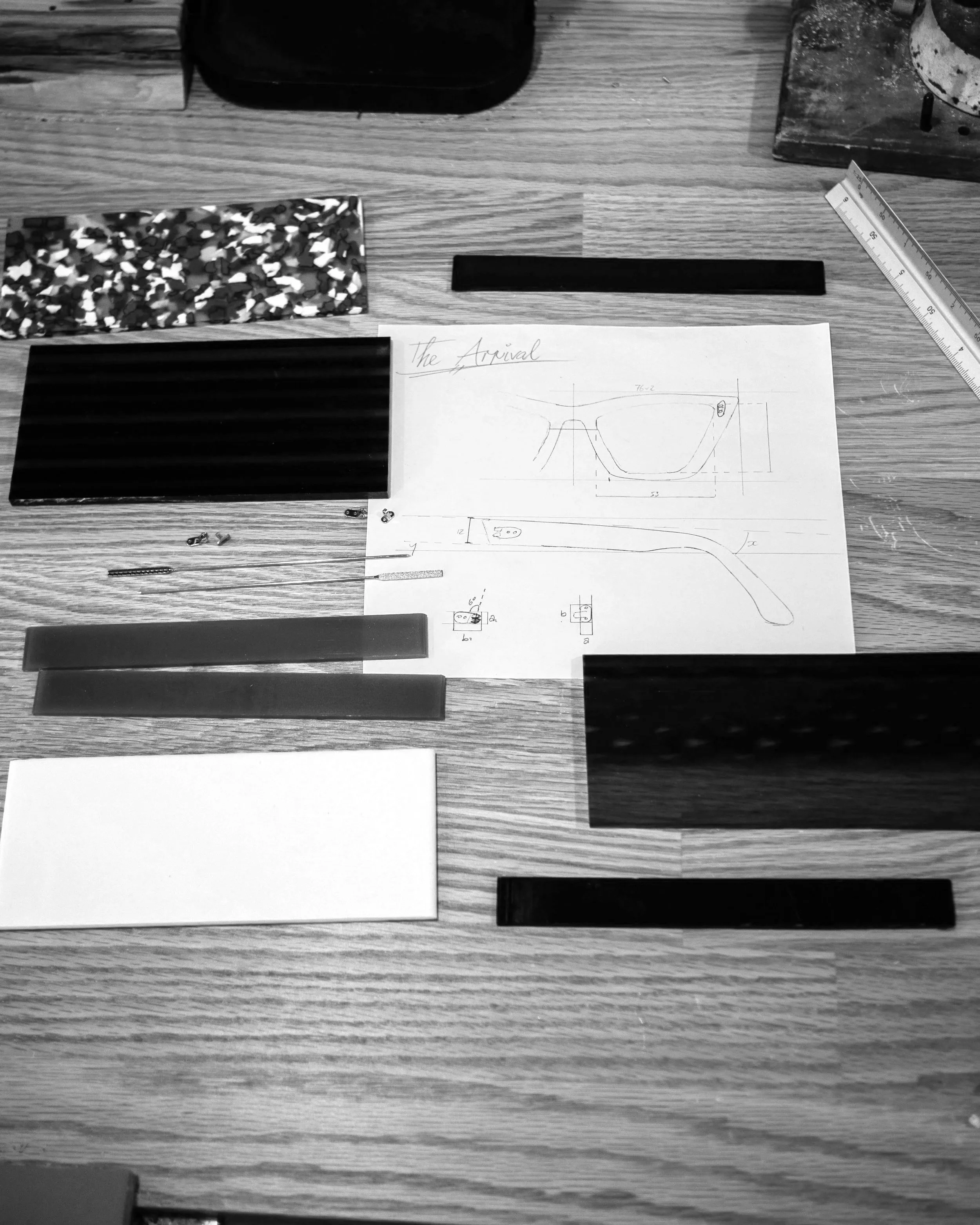 A wooden table with various objects including a ruler, a sketch of glasses labeled 'The Arrival', black and white rectangular pieces, and small metal tools.