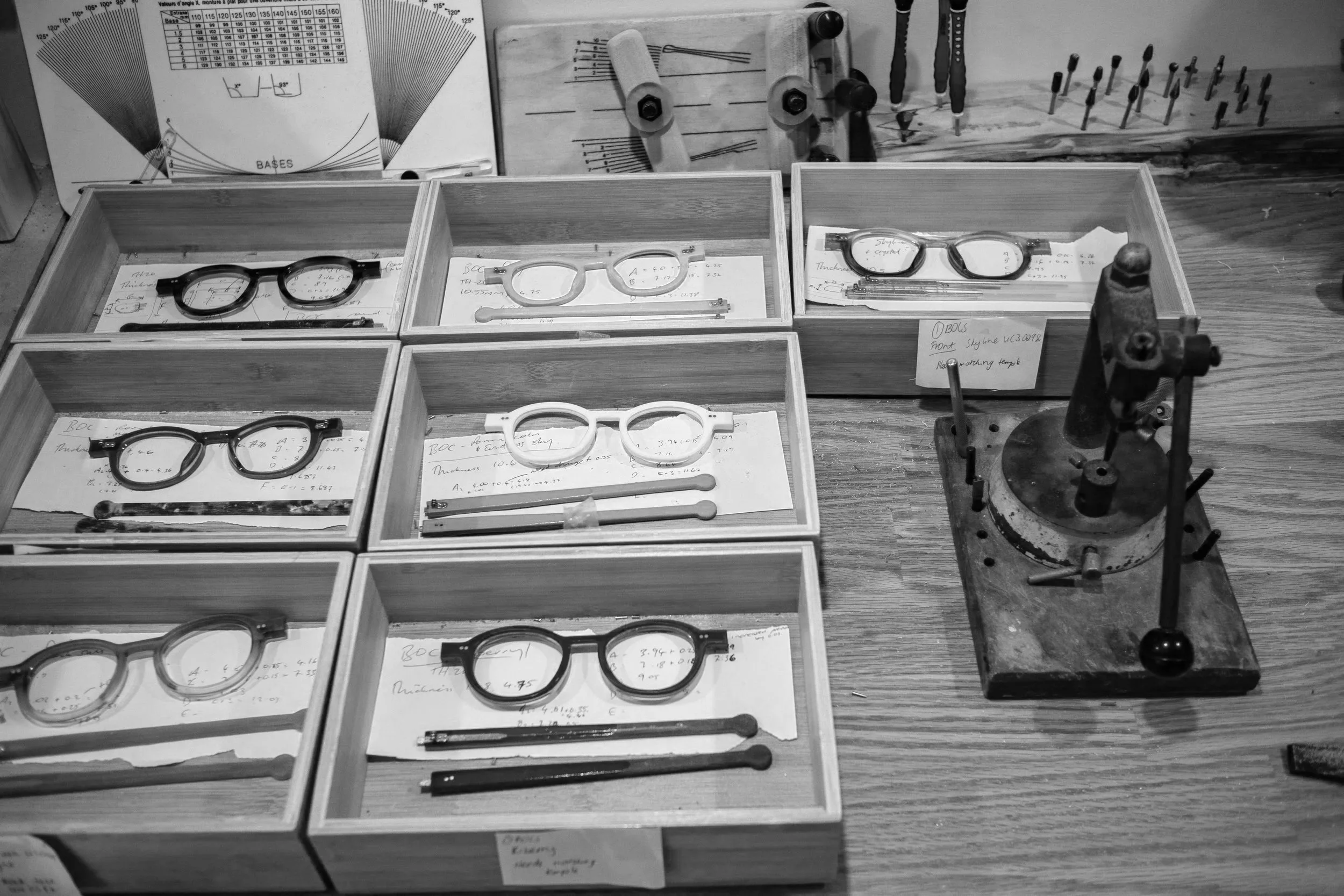 A workspace with multiple wooden boxes containing eyeglasses, mechanical parts, and handwritten notes, with tools and charts in the background.