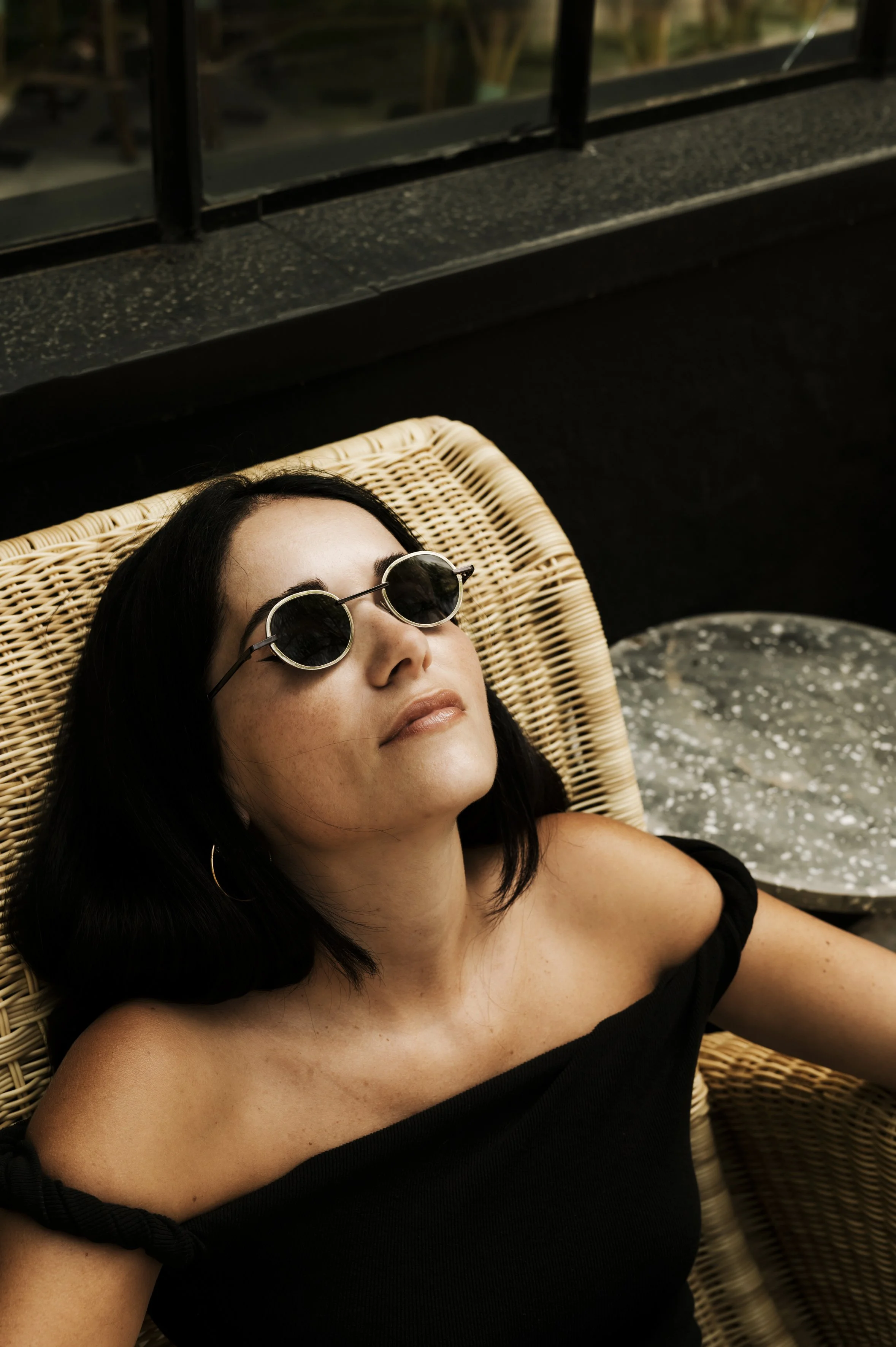 A woman with black hair and sunglasses relaxing on a wicker chair with a black off-shoulder top, near a window with a black railing, and a table with a gray marble top beside her.