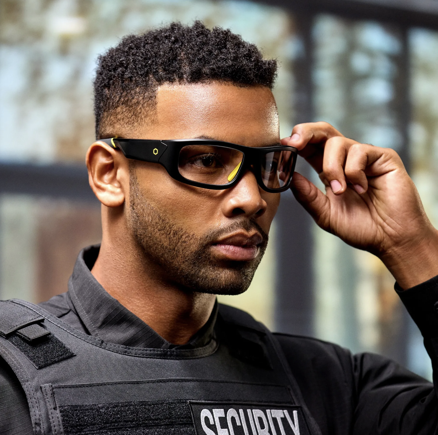 A man in a security uniform adjusting his black glasses with yellow accents, standing outdoors.