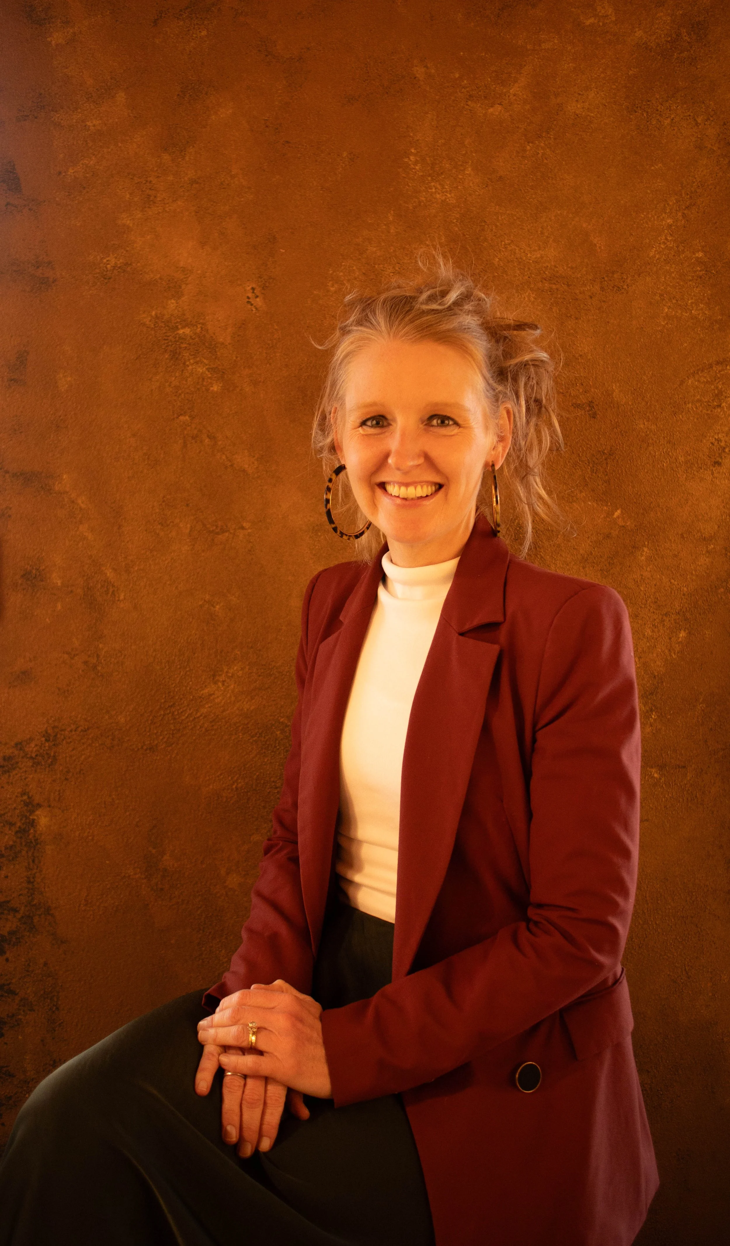 A woman with blonde hair tied back, wearing a burgundy blazer, white turtleneck, and large hoop earrings, smiling while sitting beside a textured brown and gold background.