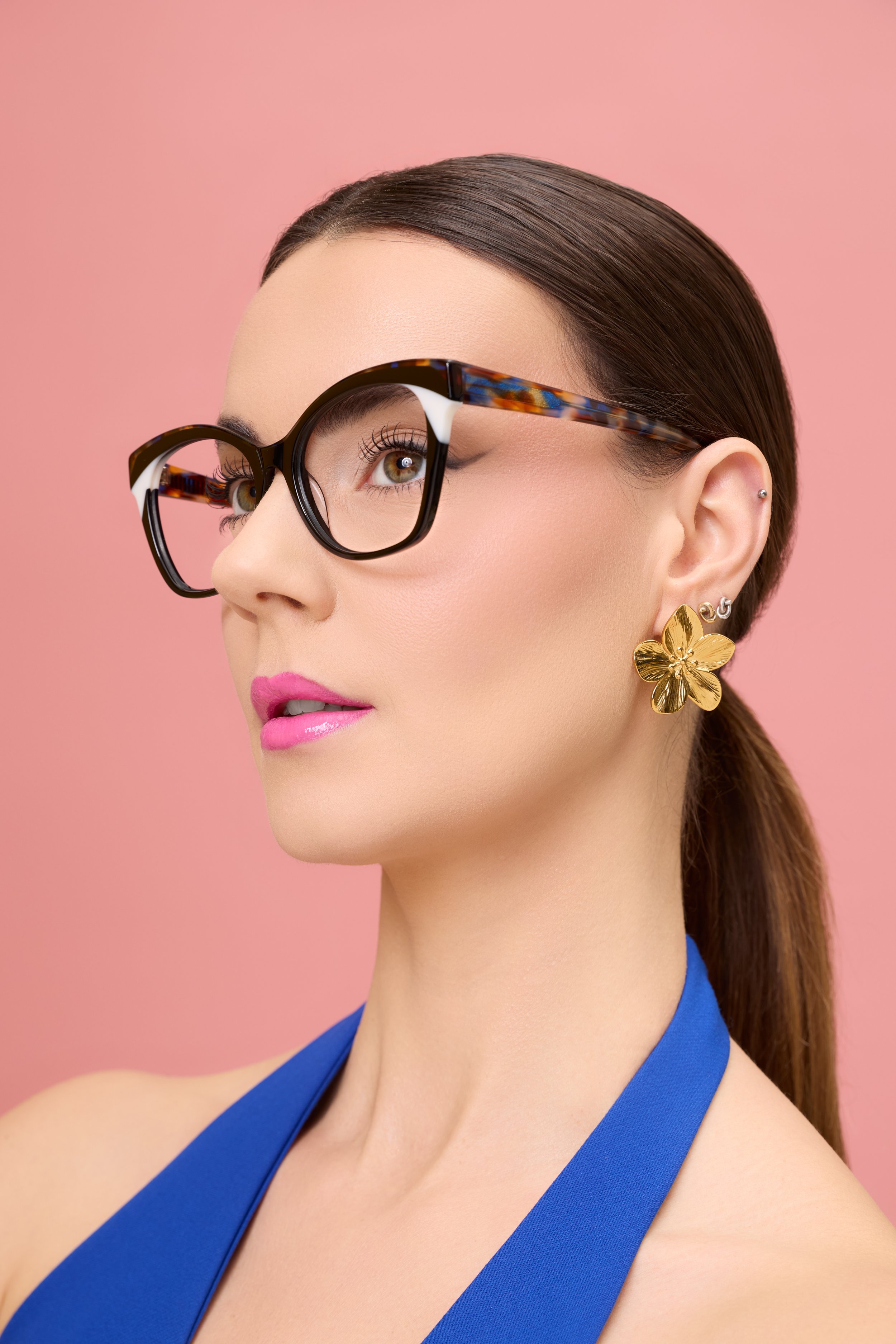 Close-up of a woman with brown hair, glasses with tortoise shell frames, gold flower earrings, pink lipstick, against a pink background, wearing a blue top.