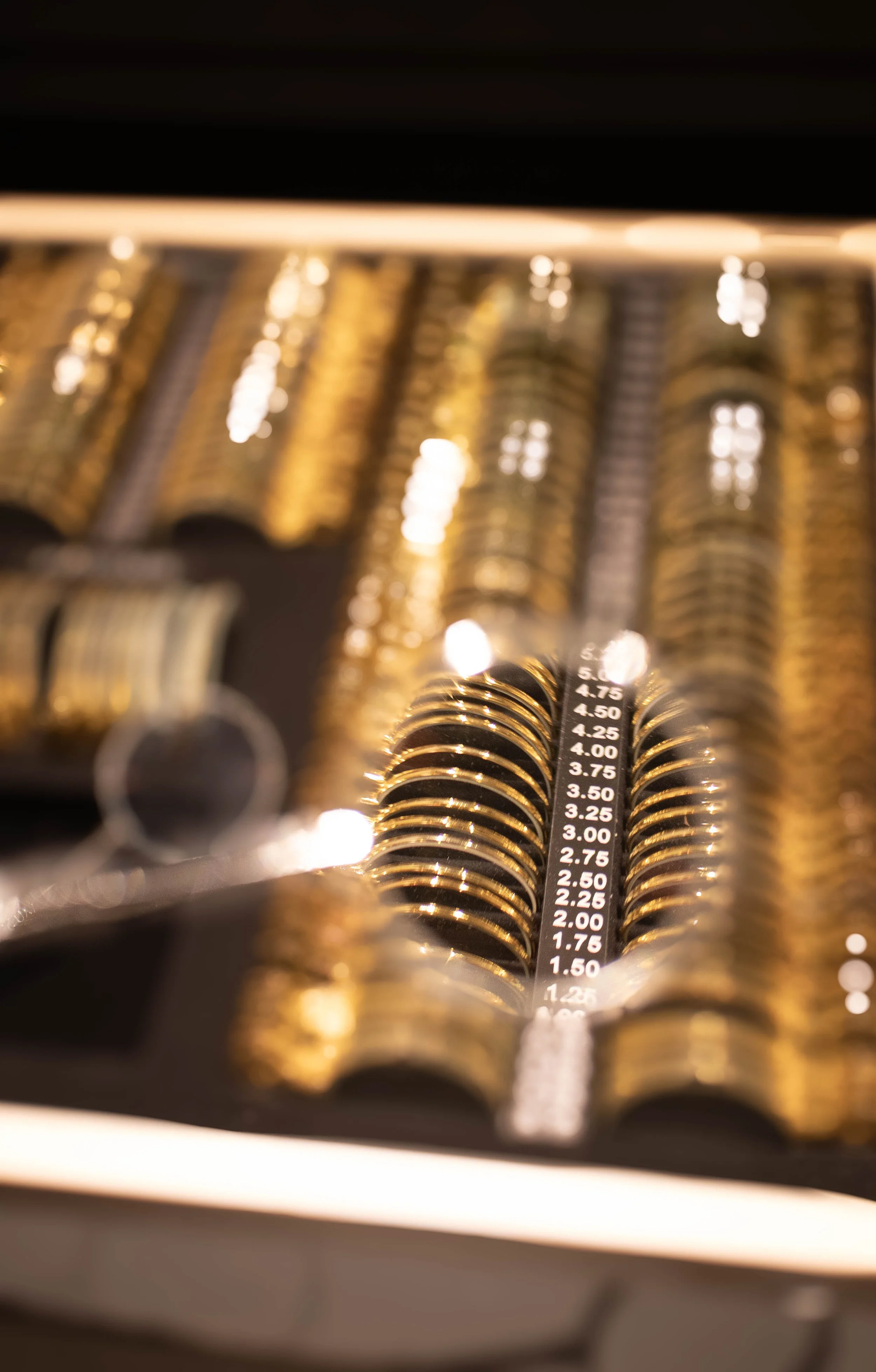 Close-up of a black measurement tool with white numbers, surrounded by blurry gold-colored objects, possibly coins or metallic rings, with bright lights reflecting on their surfaces.
