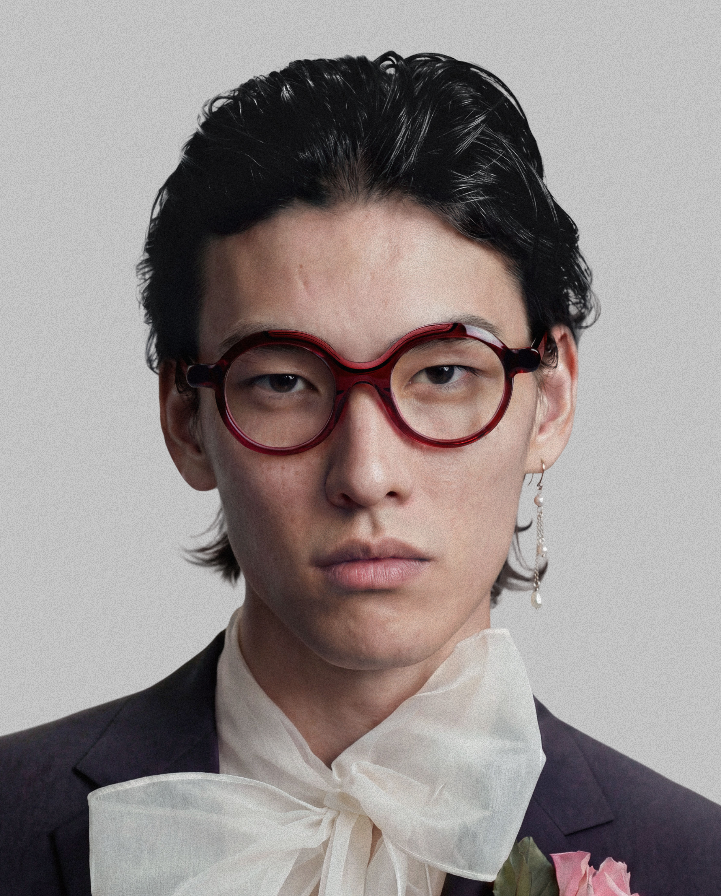 Close-up of a woman with short dark hair, wearing large round red glasses, earrings, a black suit with a white shirt, and holding pink roses.