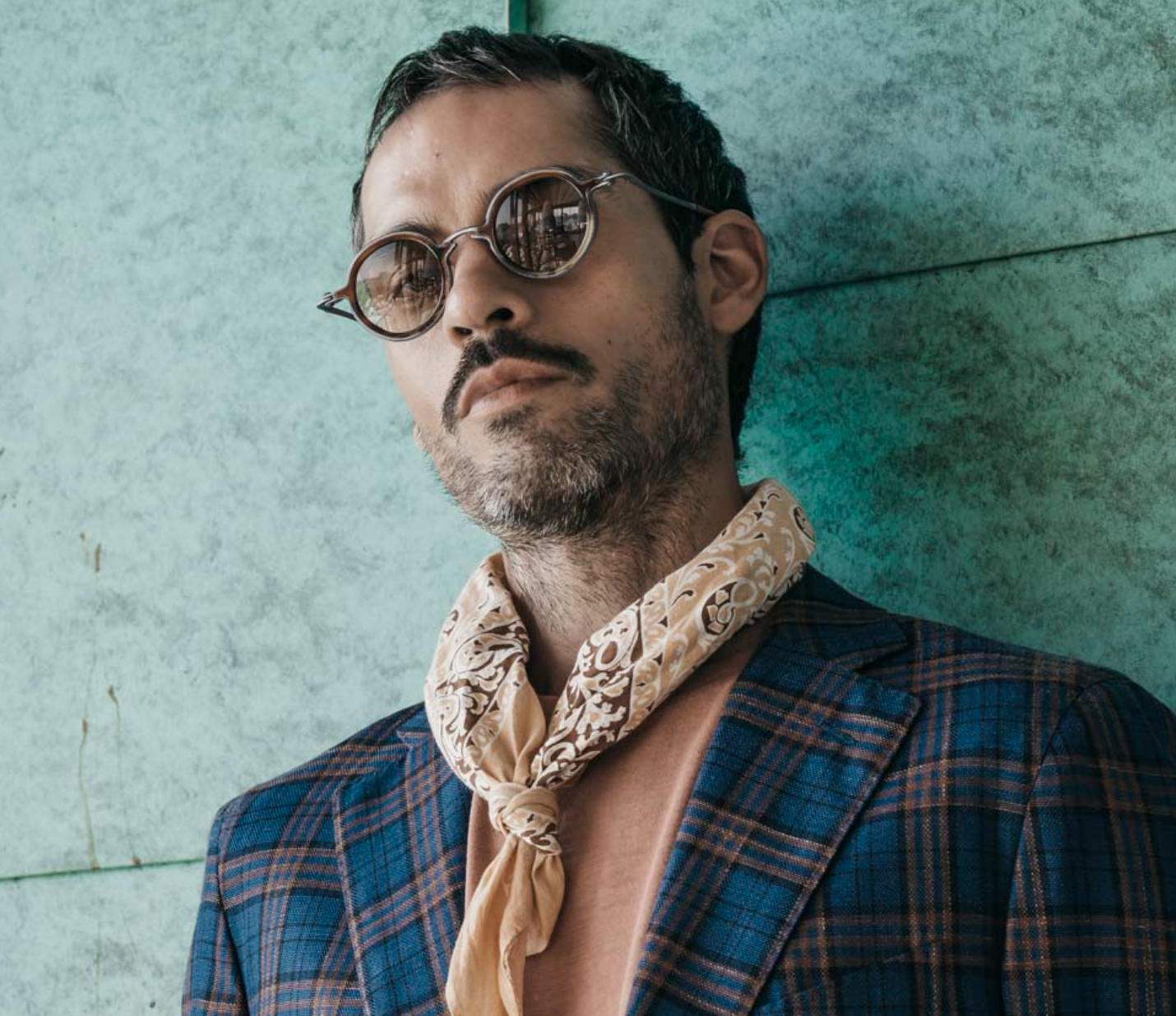 A stylish man with dark hair and a beard wears sunglasses, a beige patterned scarf, a brown shirt, and a blue plaid blazer, standing against a textured teal wall.
