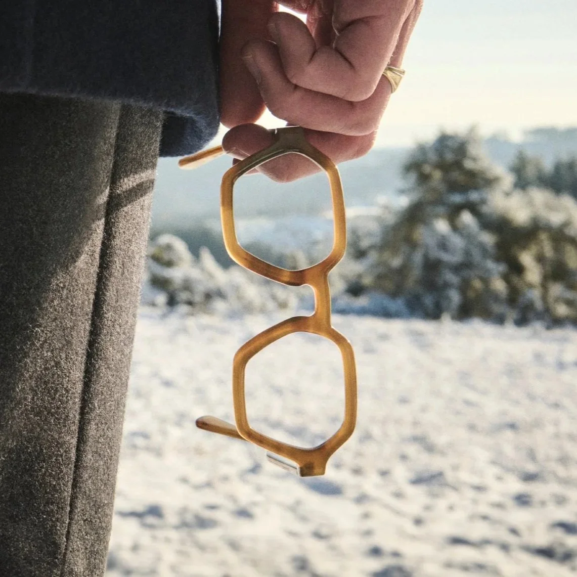 Close-up of a person's hand holding a pair of yellow sunglasses against a snowy landscape backdrop.