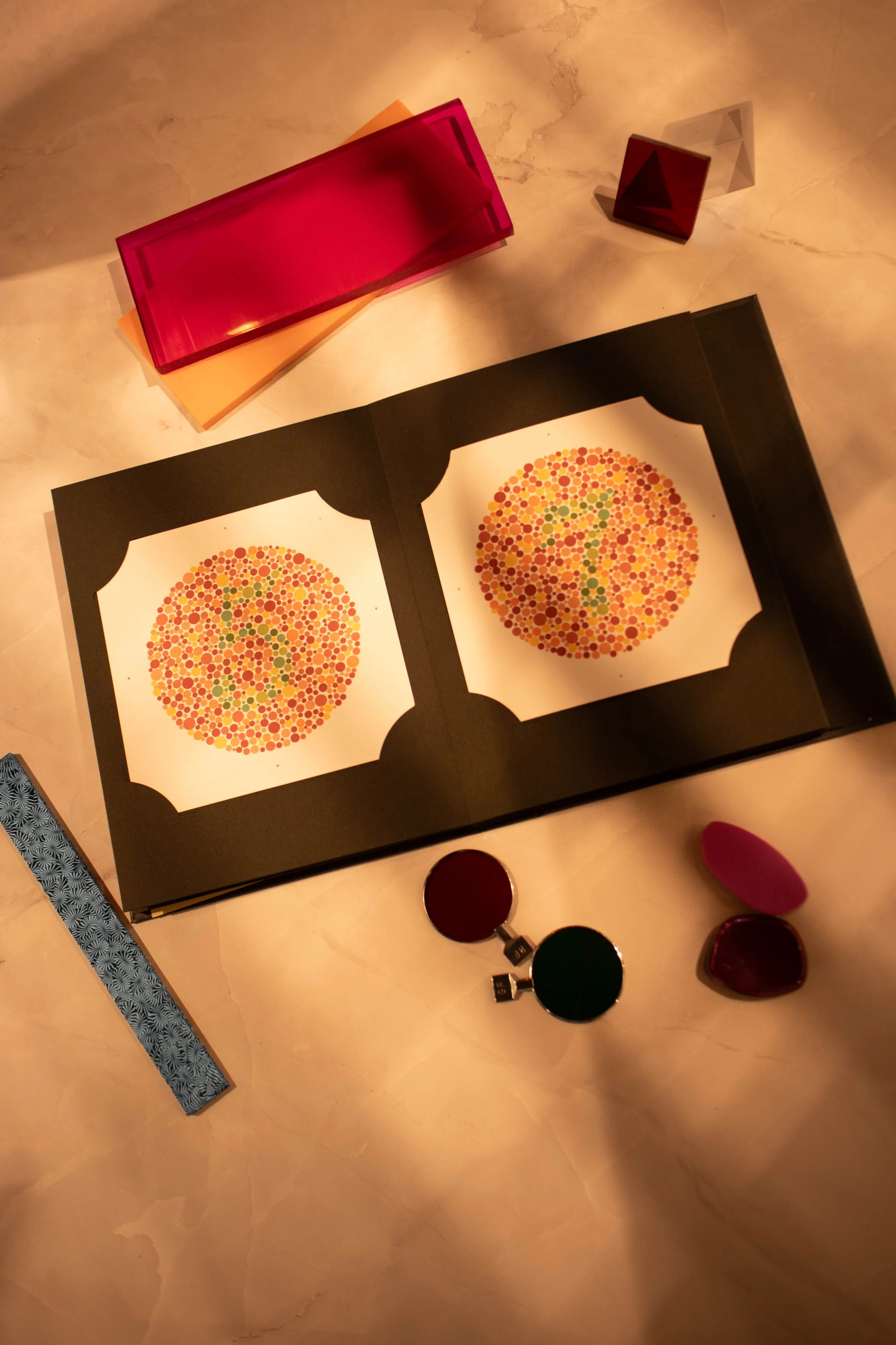 A table with colorful books, a geometric art print, and small objects including glasses of lipstick and a lighter.