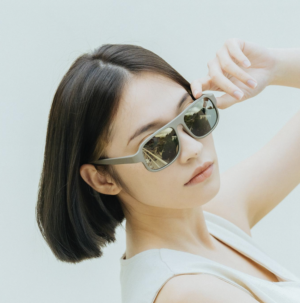 Woman with short dark hair wearing reflective sunglasses, posing with one hand near her forehead against a plain white background.