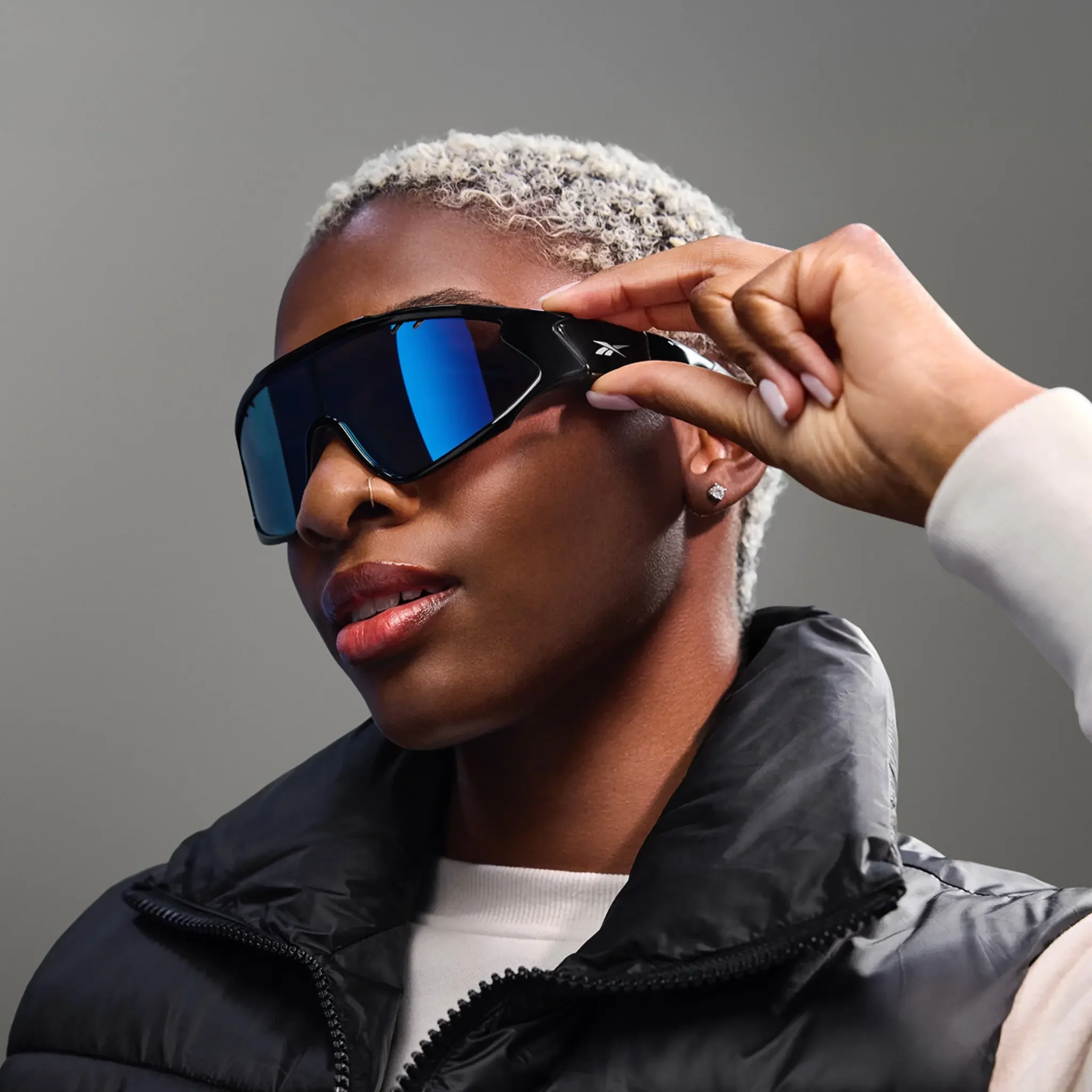 Close-up of a person with short, platinum blonde curly hair wearing large black sunglasses and a black quilted jacket, against a neutral gray background.