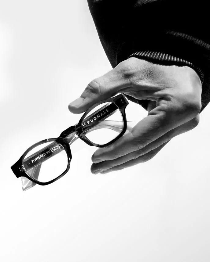 A person's hand holding a pair of eyeglasses with a black frame, against a plain background.