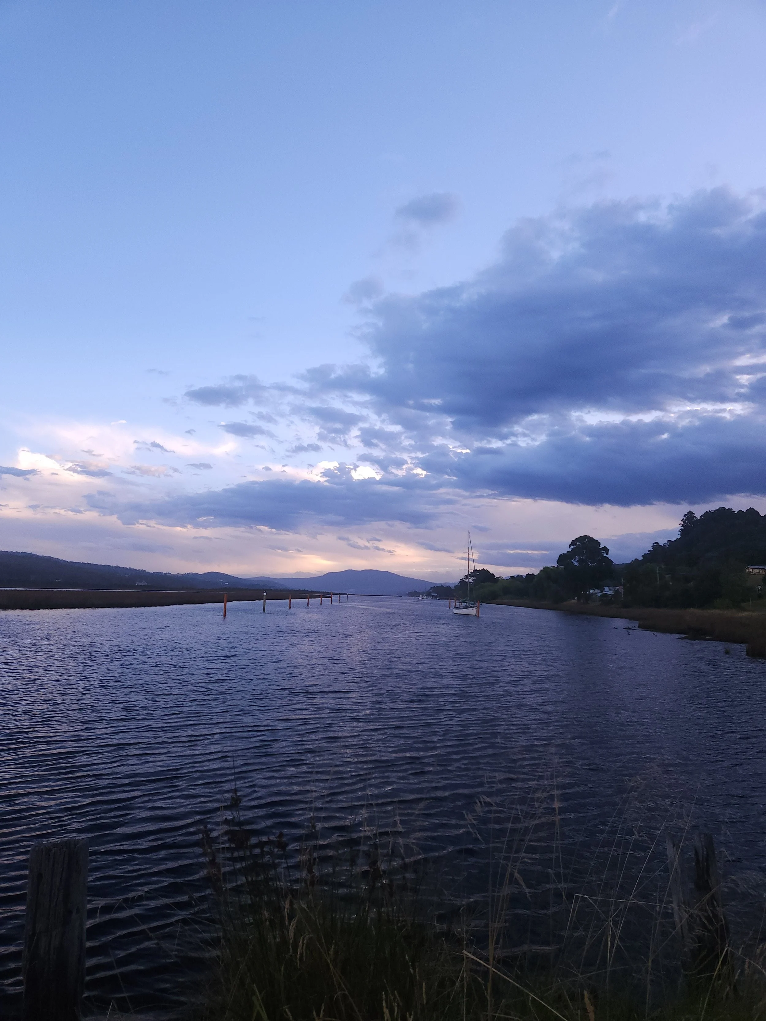 A tranquil river scene during evening with dark green trees on the right, a sailboat in the water, and mountains in the background beneath a mostly cloudy sky with hints of blue and purple.