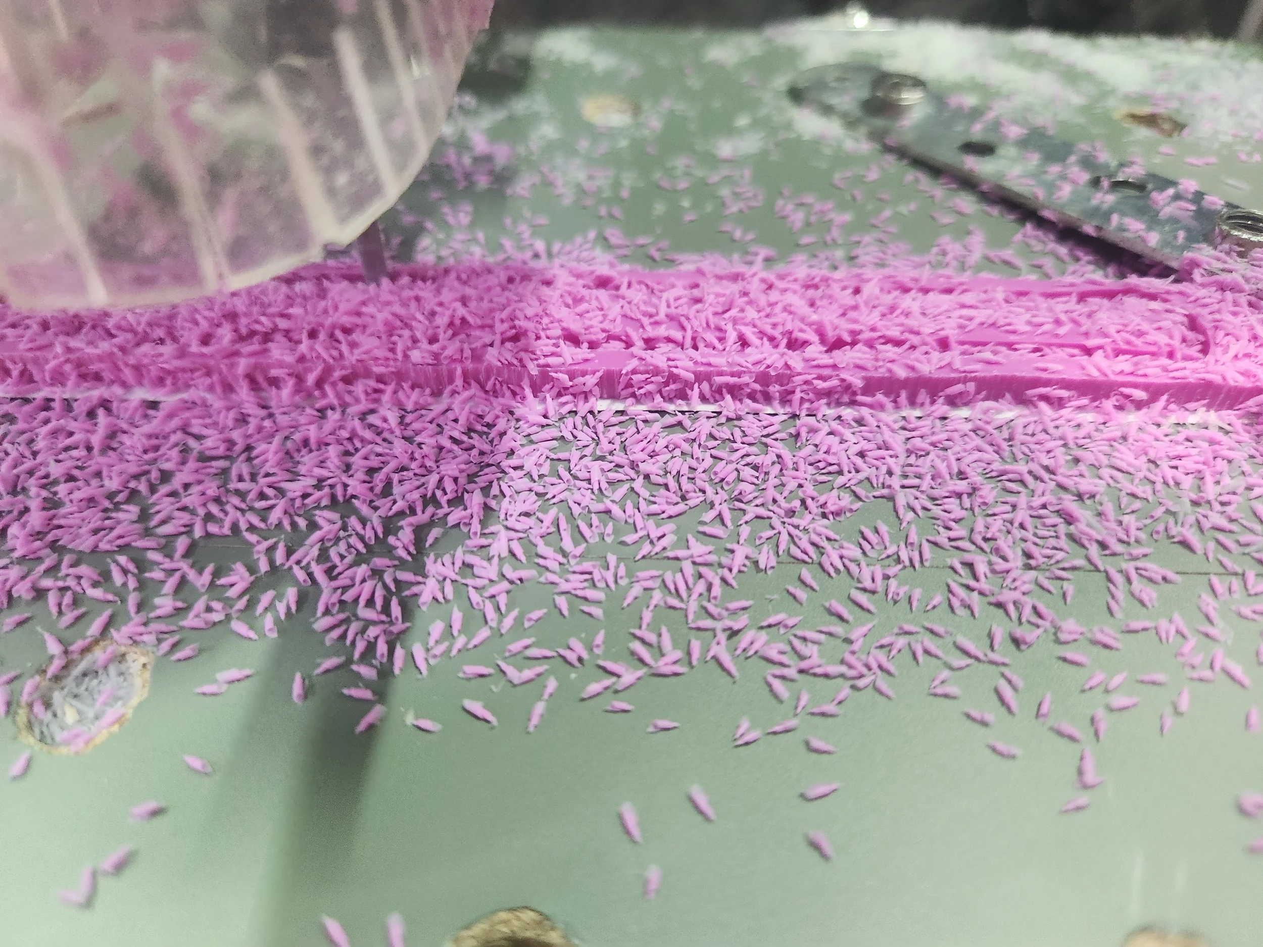 Close-up of pink shredded paper scattered on a green surface with a metallic object and part of a transparent container nearby.