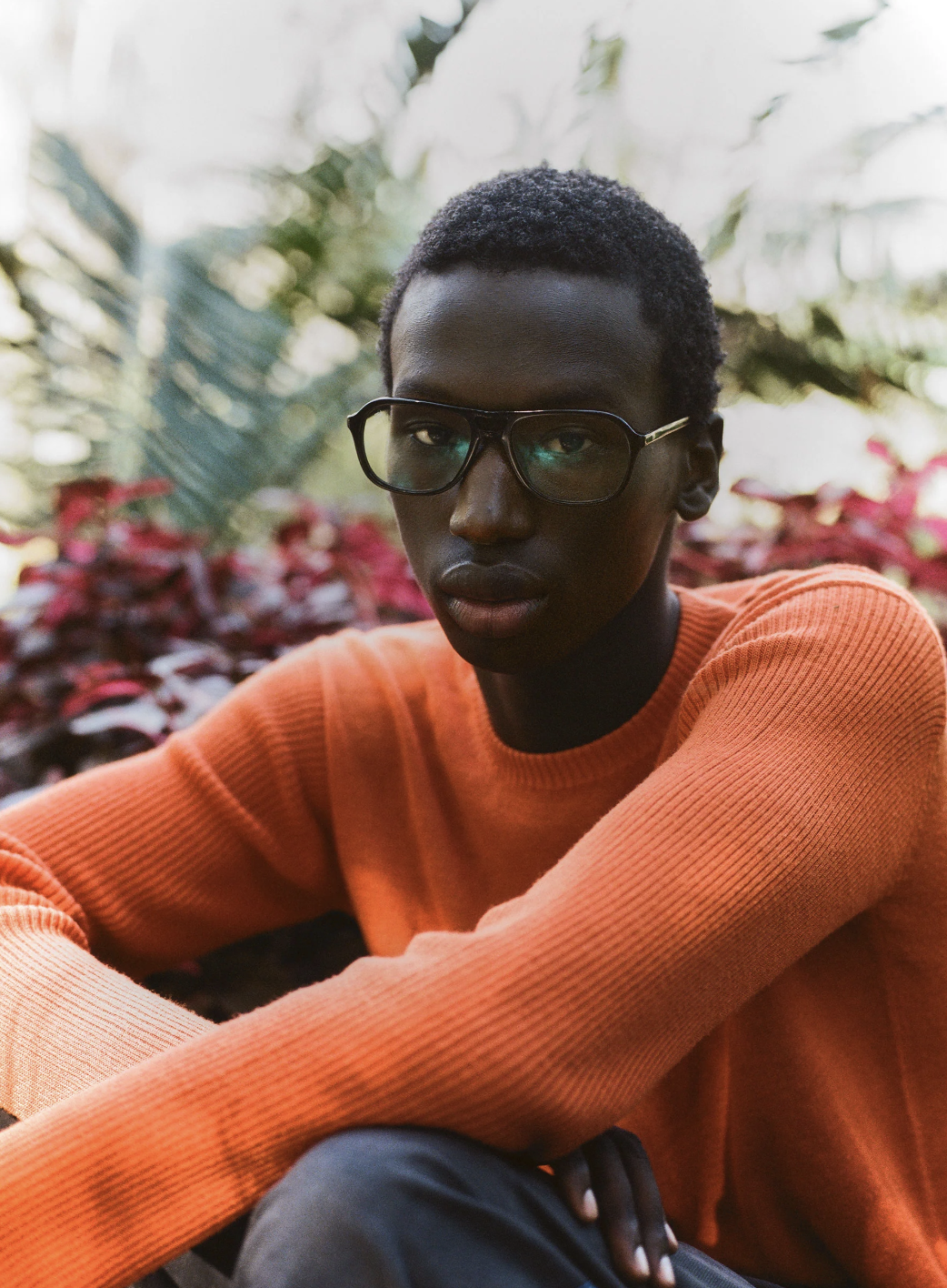 A young man with dark skin, short curly hair, and glasses, wearing an orange sweater, sitting outdoors with a background of blurred greenery and red foliage.