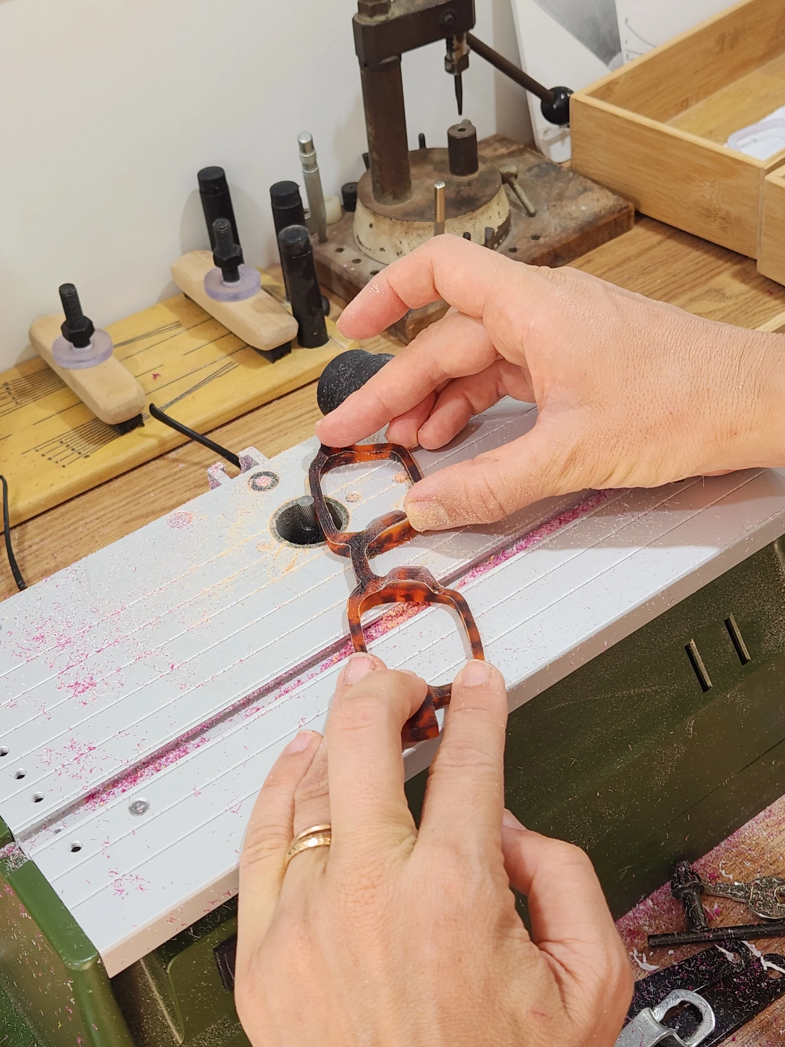 Close-up of hands working on a gasket with equipment in a workshop or garage. The person is using a tool to align a gasket on a work surface, with a wooden workbench holding various machinery and tools in the background.