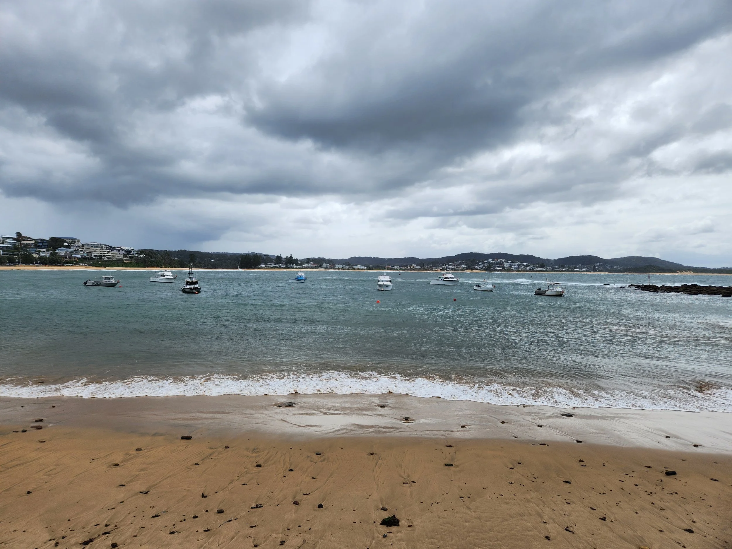 A beach with light brown sand, scattered with small rocks and seaweed, facing a body of water with small motorboats anchored near the shore. In the background, there are residential houses along the coastline and rolling hills under a cloudy, overcast sky.
