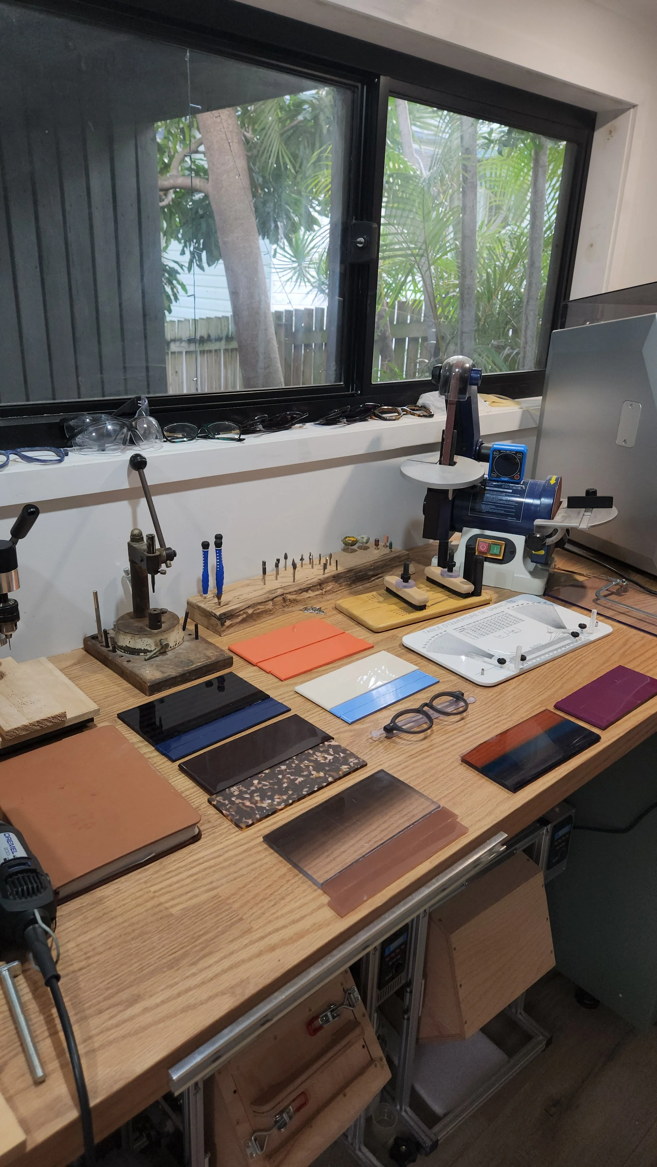 Workstation with precision tools, sample tiles, and glasses, set near a window with a view of trees and a wooden fence.