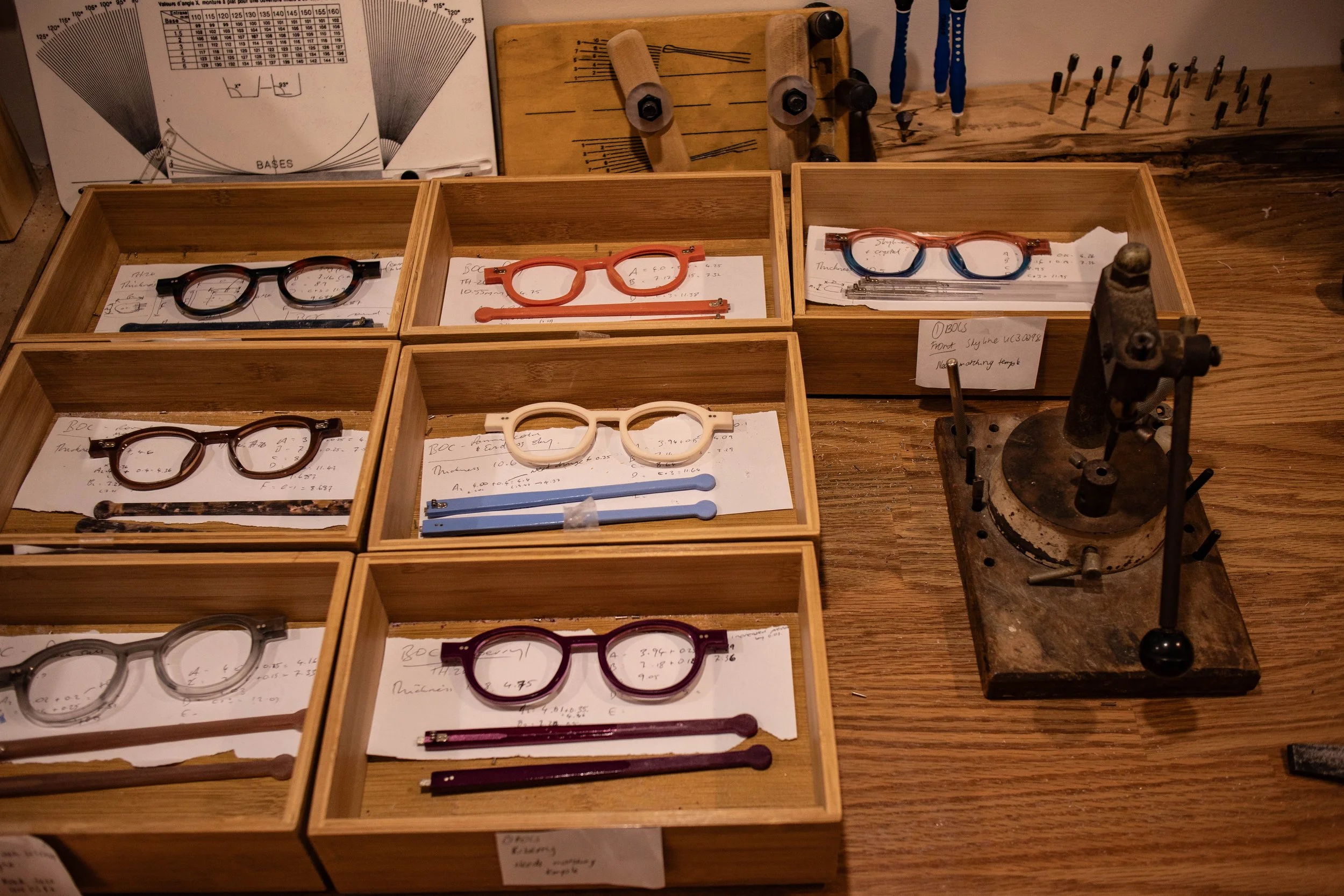A workbench with wooden boxes containing eyeglasses and writing tools, and an assortment of tools and charts related to optical or eyewear work.