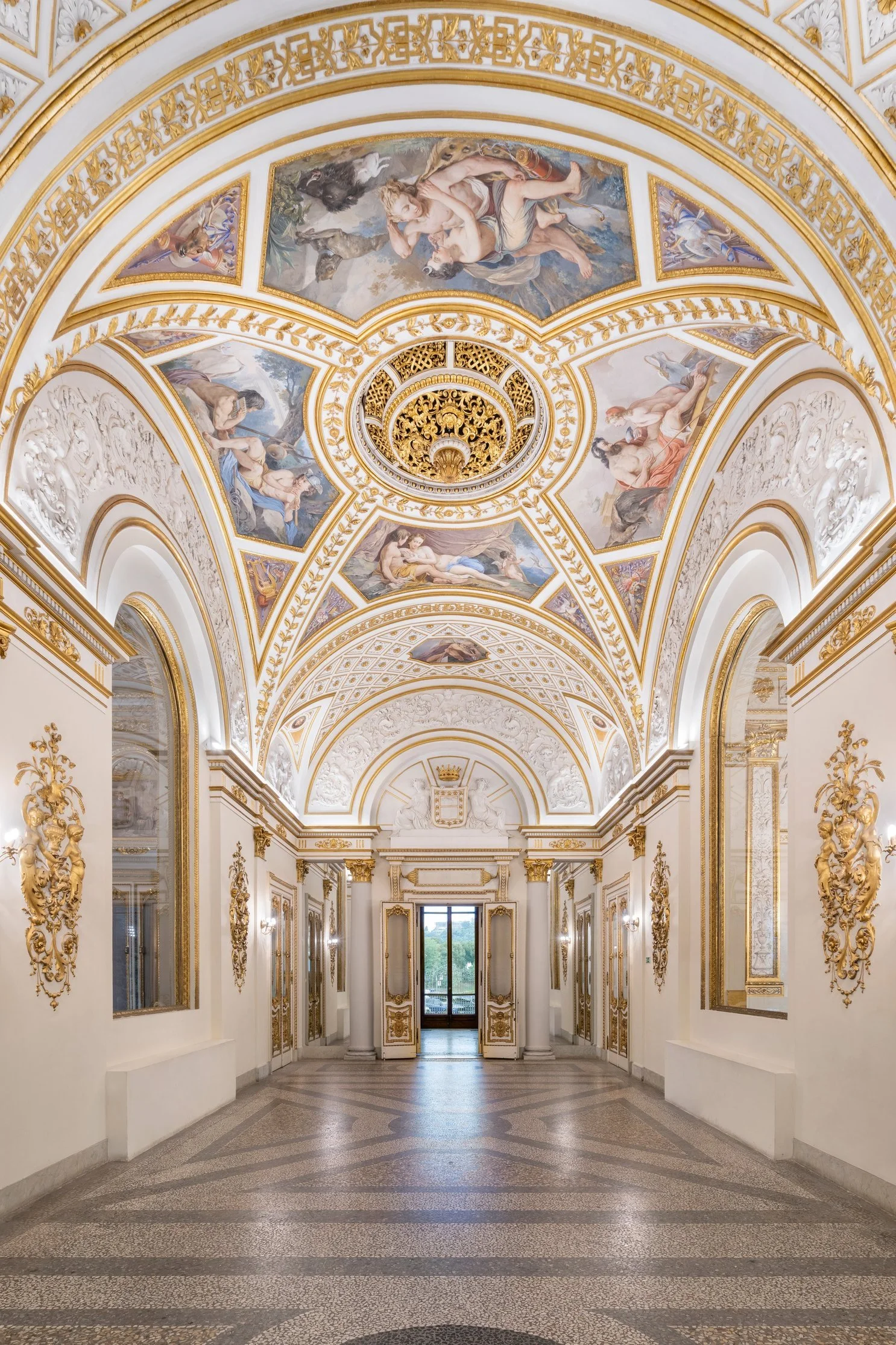 Elegant hallway with ornate gold and white decorations, detailed ceiling paintings of mythological scenes, tall columns, and large windows at the end providing natural light.