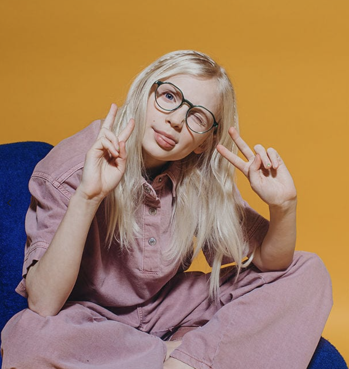 A young woman with long blonde hair, wearing glasses and a pink outfit, making peace signs with both hands against a yellow background.