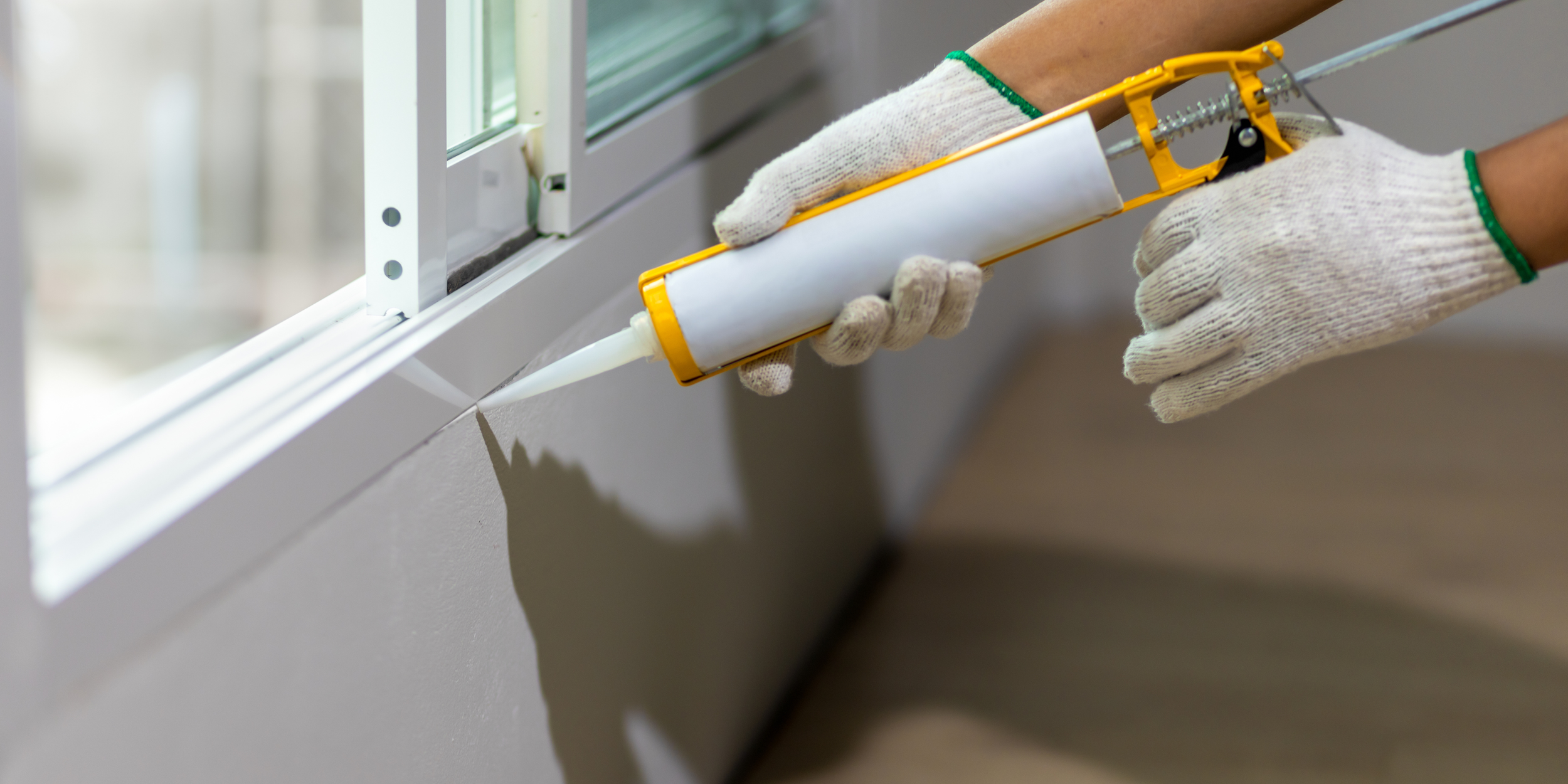 A person wearing white gloves applying white caulk along the edge of window trim with a caulking gun.