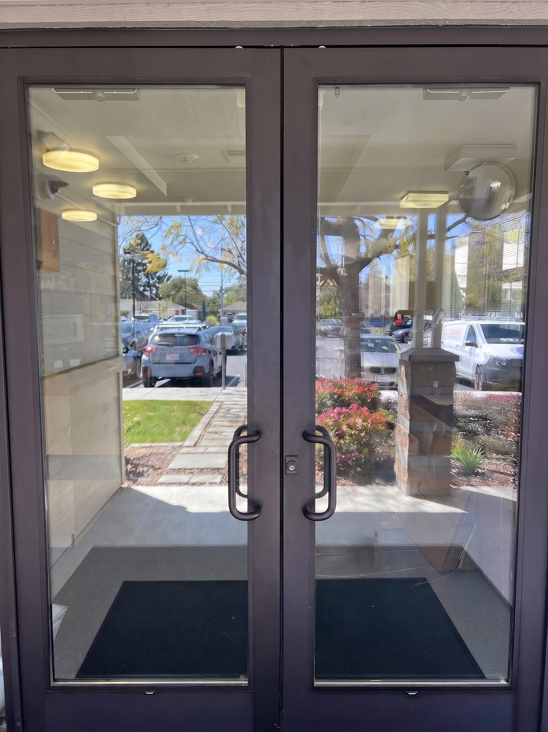 Double glass doors with black metal frames, reflecting parking lot and outside trees, with black rubber mats inside.