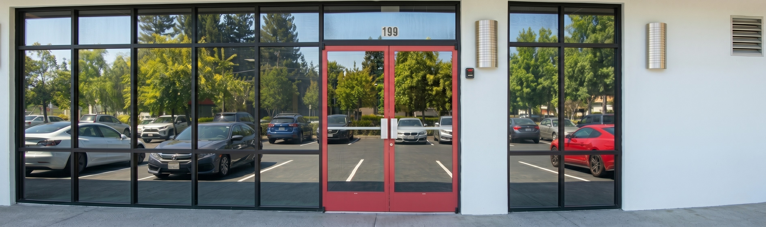 Front view of a commercial building with large glass windows reflecting trees and parking lot, with a red door at the center.