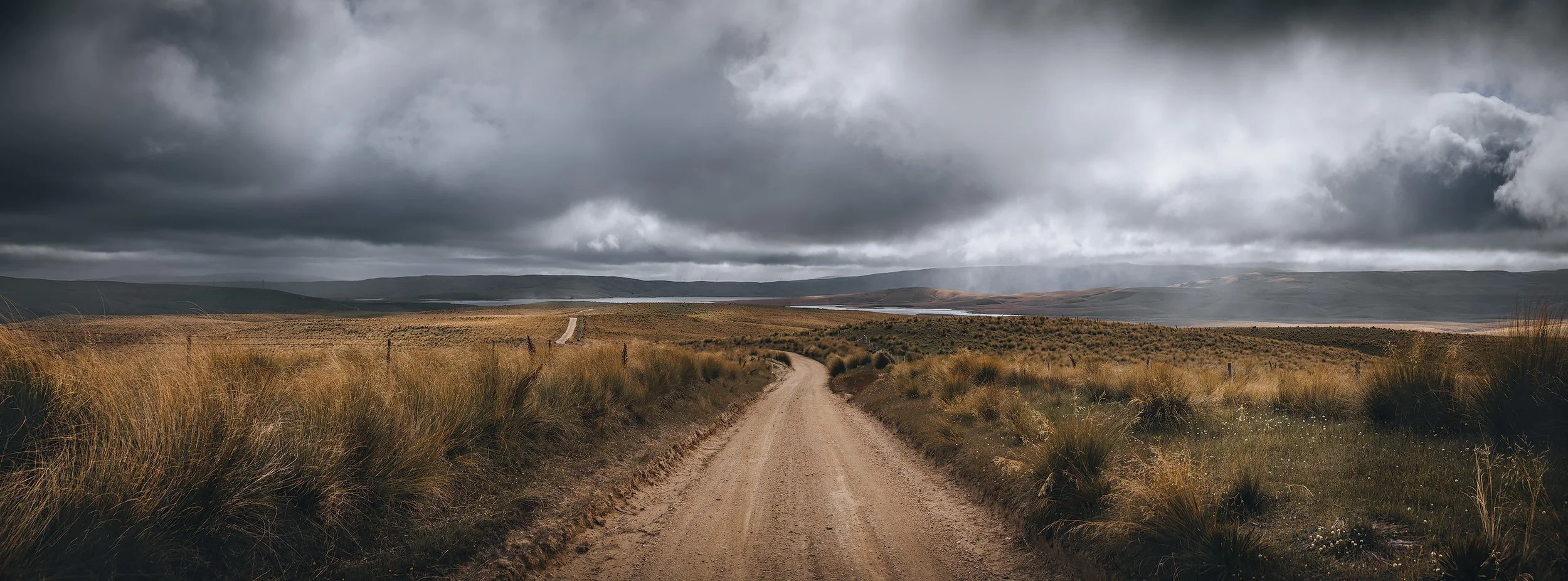 Panoramic high country landscape with a gravel road winding through golden tussock beneath dramatic storm clouds in New Zealand.