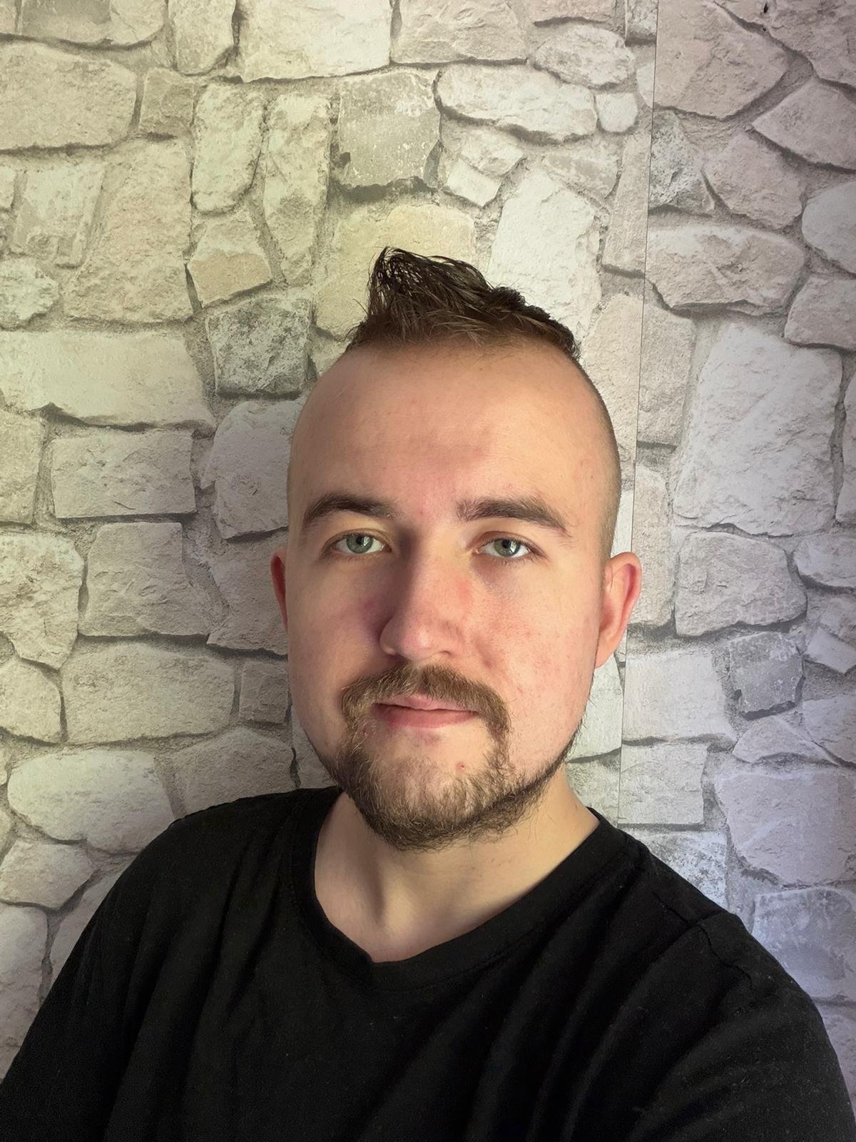 Ein junger Mann mit kurzen Haaren, Vollbart und einem schwarzen T-Shirt, der vor einer Steinmauer fotografiert wurde.