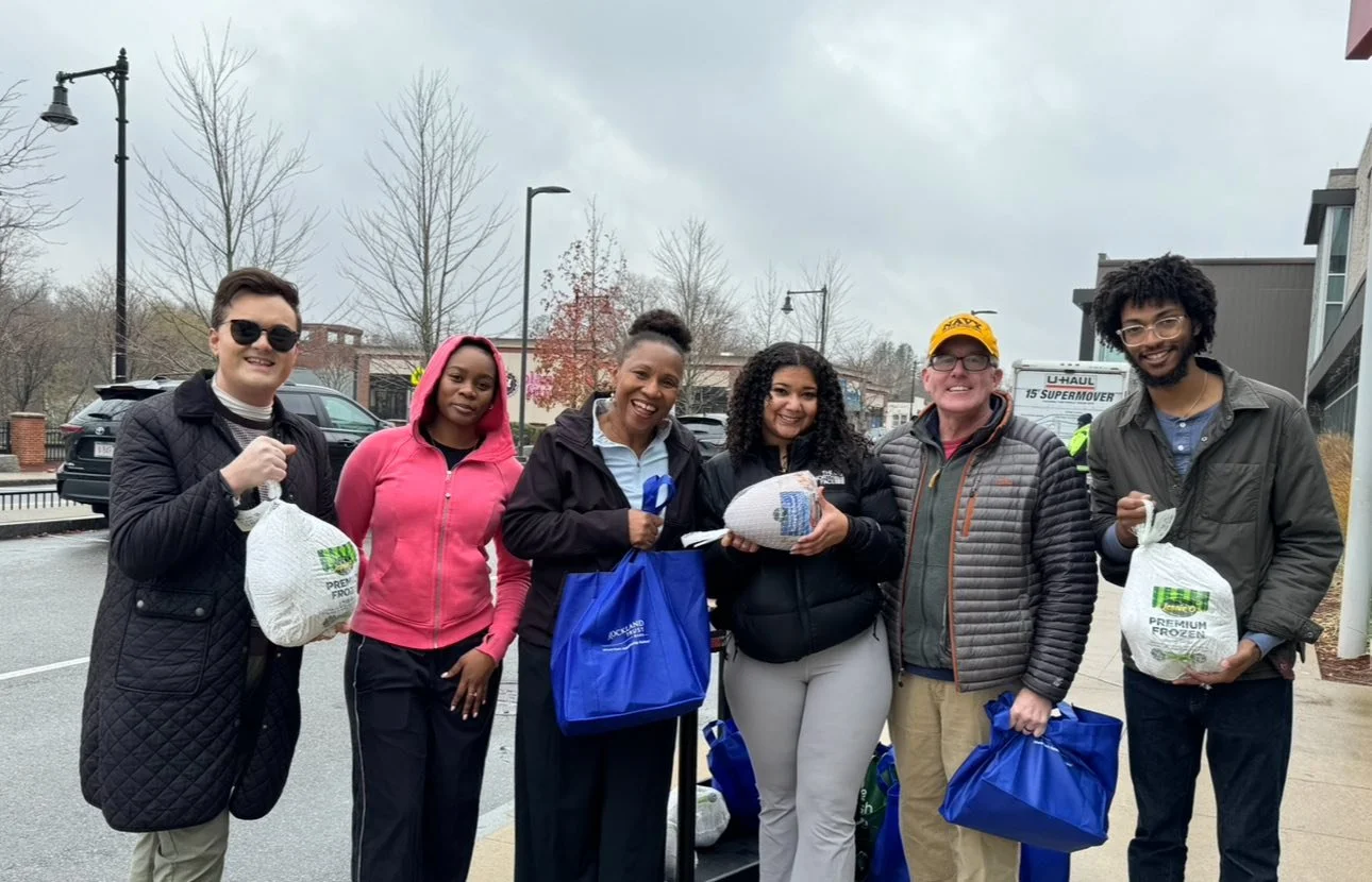 Group of six diverse people standing outdoors, holding bags of frozen food and smiling, on a cloudy day in a parking lot.