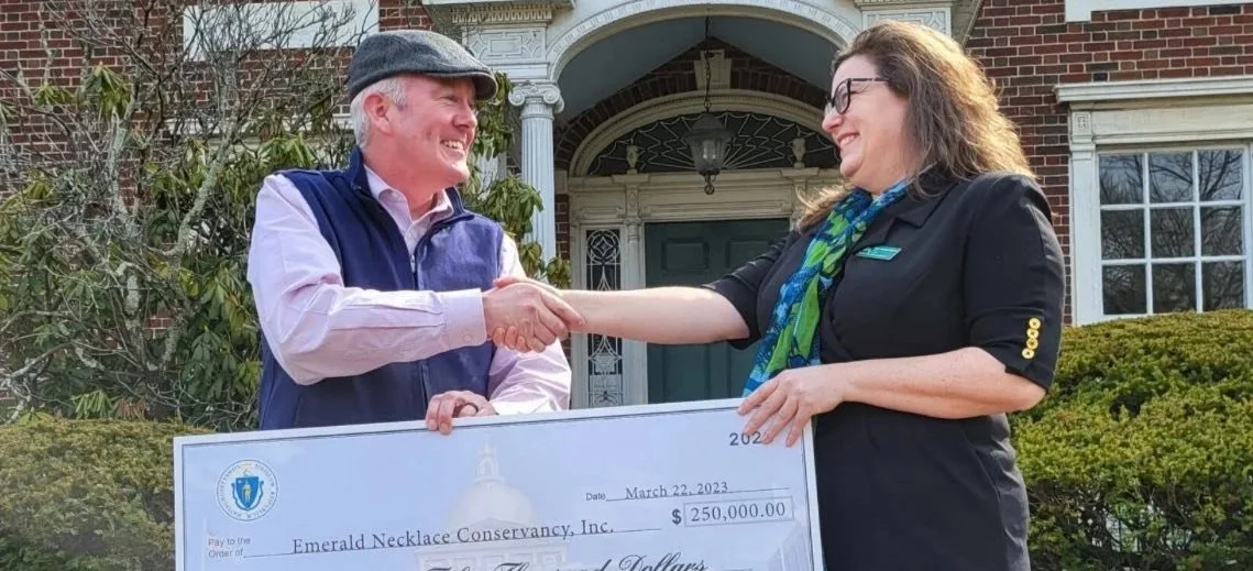 A woman is shaking hands with a man while holding a large check for 250,000 dollars in front of a house with brick and white trim. The woman is smiling and wearing glasses, a black blazer, and a colorful scarf. The man is smiling, wearing a cap, a purple vest over a light shirt, and is also smiling.
