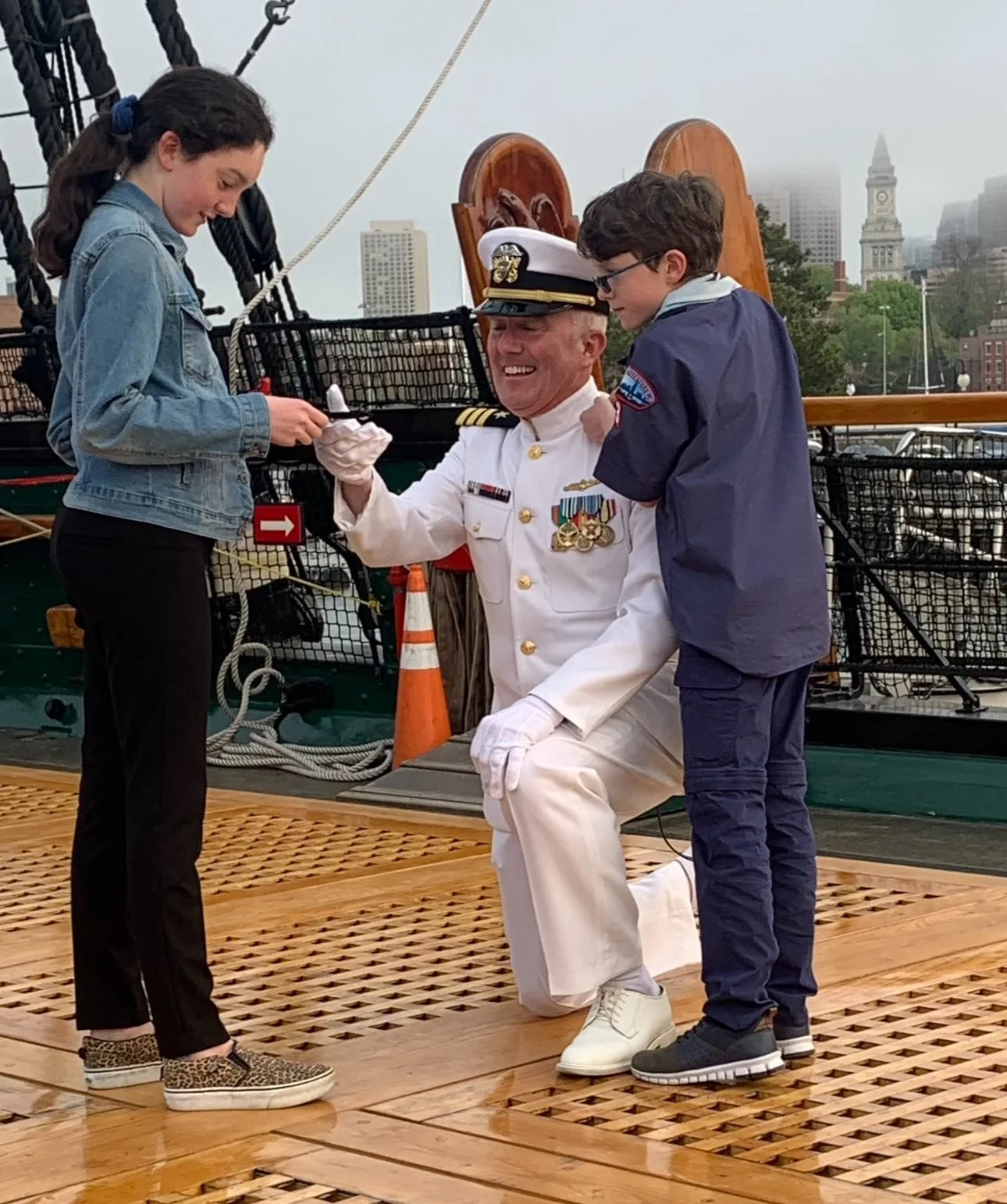 A man in a white Navy uniform with medals on his chest is kneeling on a wooden platform, smiling, as he shakes hands with a young woman in a denim jacket and black pants. Another young man in a blue jacket is-standing nearby, watching the handshake. The background shows a harbor with boats and a cityscape with tall buildings and a clock tower.