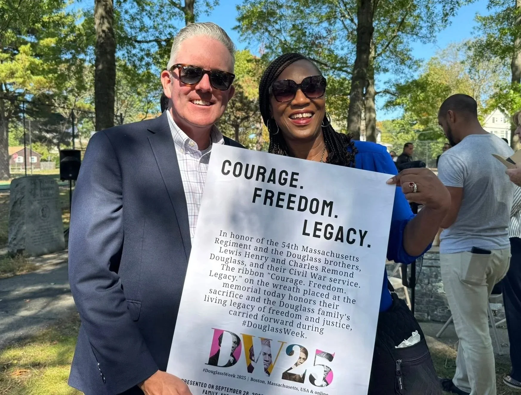 A man and woman smiling outdoors during a Civil War service commemorative event, holding a sign with text about the 54th Massachusetts Regiment and Douglass family. The man is wearing sunglasses and a suit, and the woman is wearing sunglasses and a blue jacket. There are other people and trees in the background.