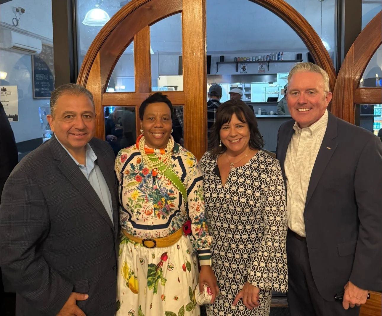 Four people smiling and posing for a photo in a restaurant. The group includes two men and two women dressed in business and colorful attire. Behind them is a wooden window frame and a busy kitchen area.