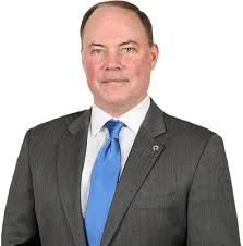 Portrait of a man in a suit and blue tie, smiling against a white background.