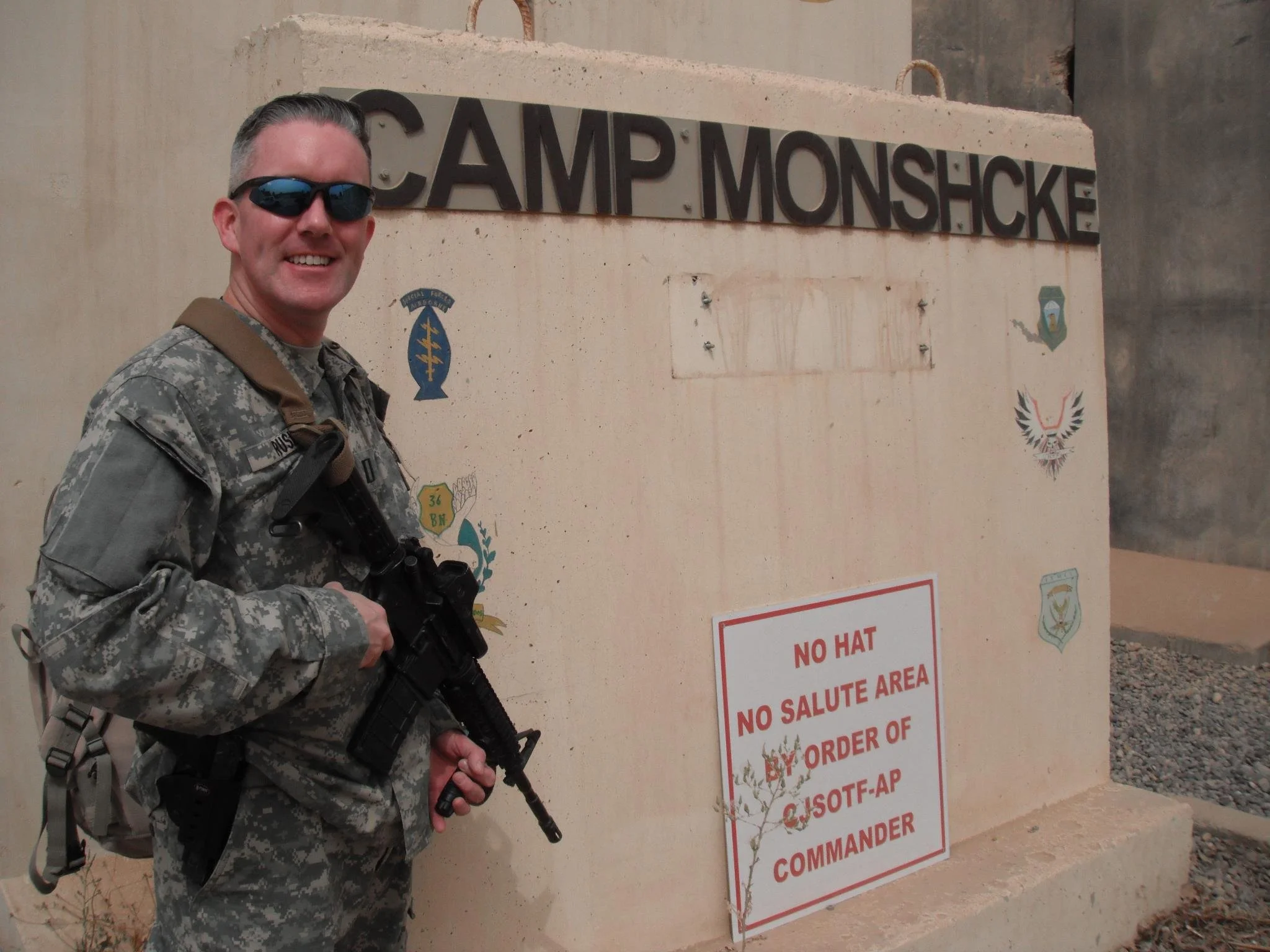A smiling man in military camouflage uniform, wearing sunglasses and carrying a rifle, stands next to a sign that reads 'No Hat, No Salute Area.' The sign is mounted on a concrete wall with various military insignias and symbols.
