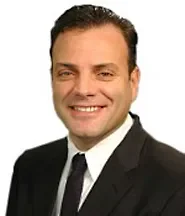 A man with dark hair, wearing a black suit, white shirt, and black tie, smiling against a white background.