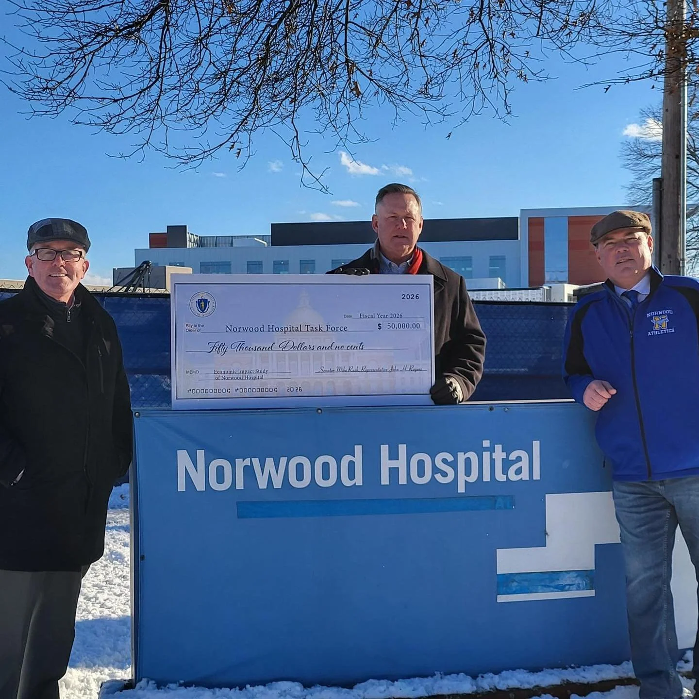 Three men standing outdoors in winter, holding a large check for $50,000 made out to Norwood Hospital, with a hospital building in the background.