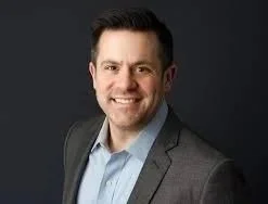 A man in a gray suit smiling against a dark background.