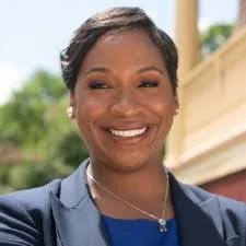 Portrait of a smiling woman wearing a navy blazer and a necklace, outdoors with greenery and a building in the background.