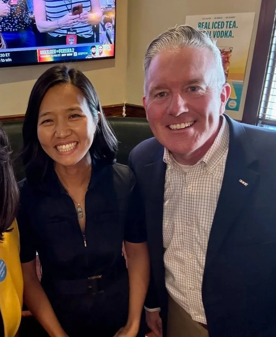 A smiling Asian woman and a smiling white man in a suit jacket posing together in a restaurant or bar, with a TV screen displaying sports and an advertisement for iced tea and vodka in the background.