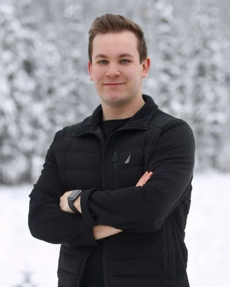 Young man standing outdoors in a snowy landscape with trees in the background, wearing a black jacket and smart watch, with arms crossed and smiling slightly.